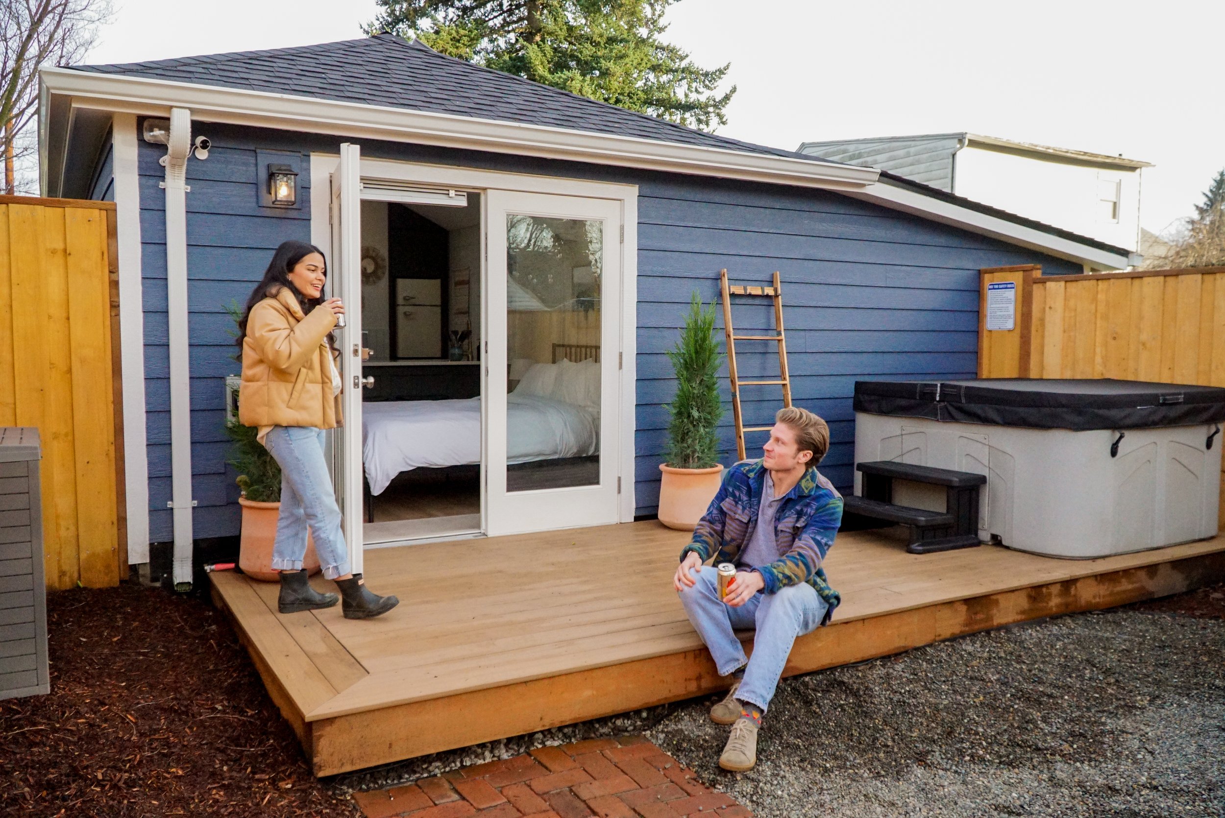 A woman standing on a wooden deck outside a blue house talking to a seated man holding a drink. The deck includes potted plants, a ladder, and a hot tub.
