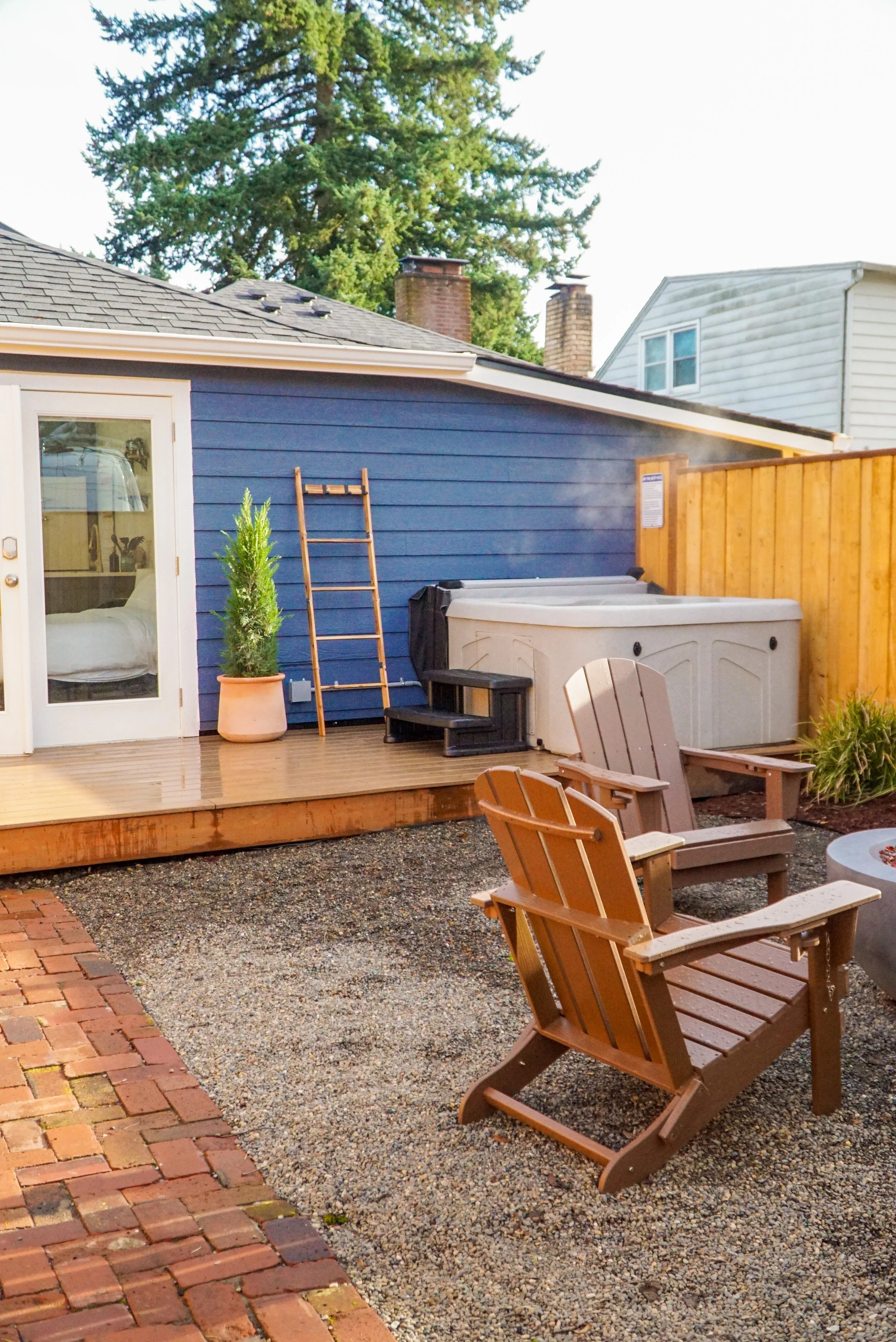 An outdoor backyard scene with a deck, two Adirondack chairs, a hot tub, a wooden ladder, potted plant, and a blue house wall with white door and glass window. A wooden fence is on the right side.