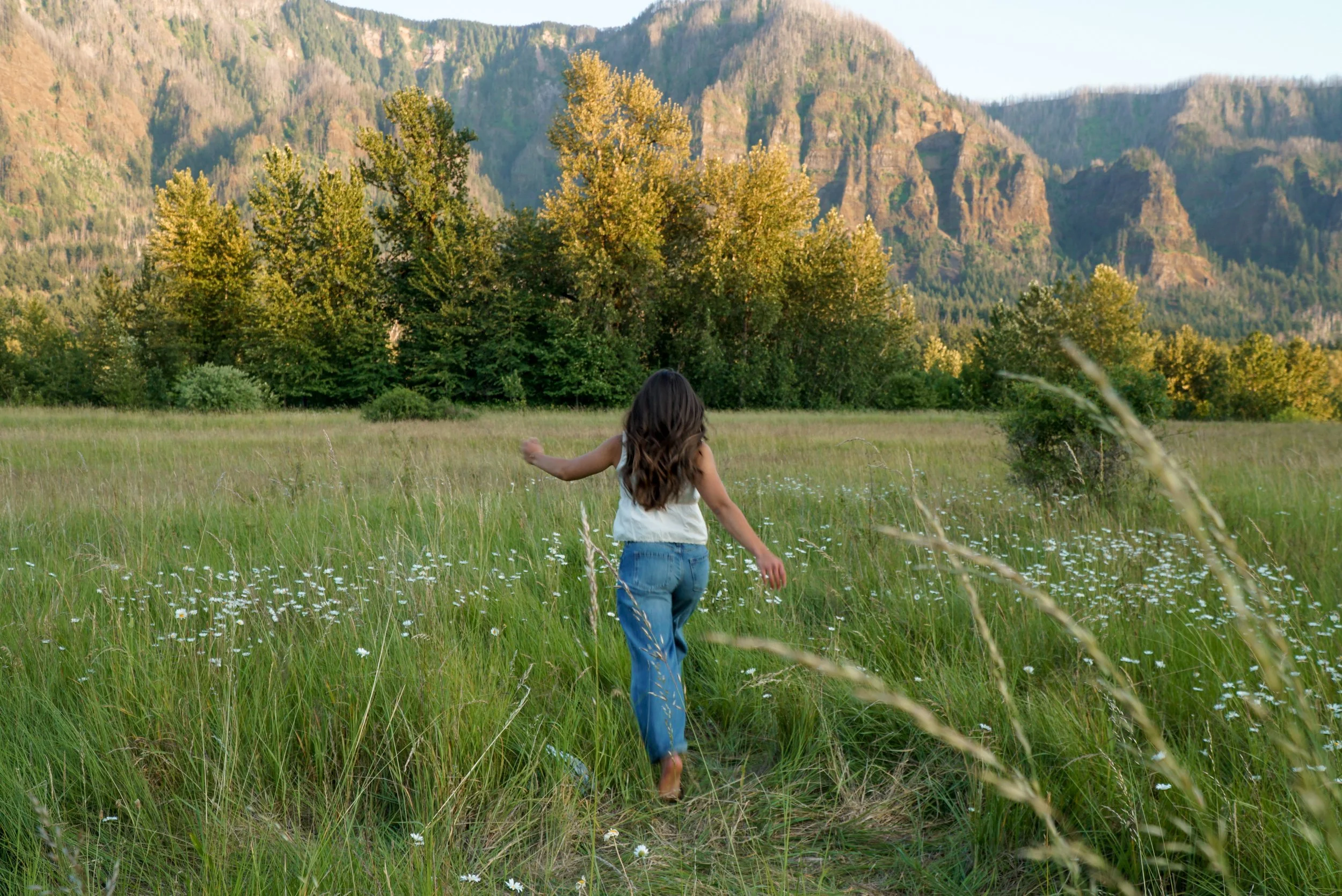 A girl walking through a grassy field with small white flowers, surrounded by trees and mountains in the background.