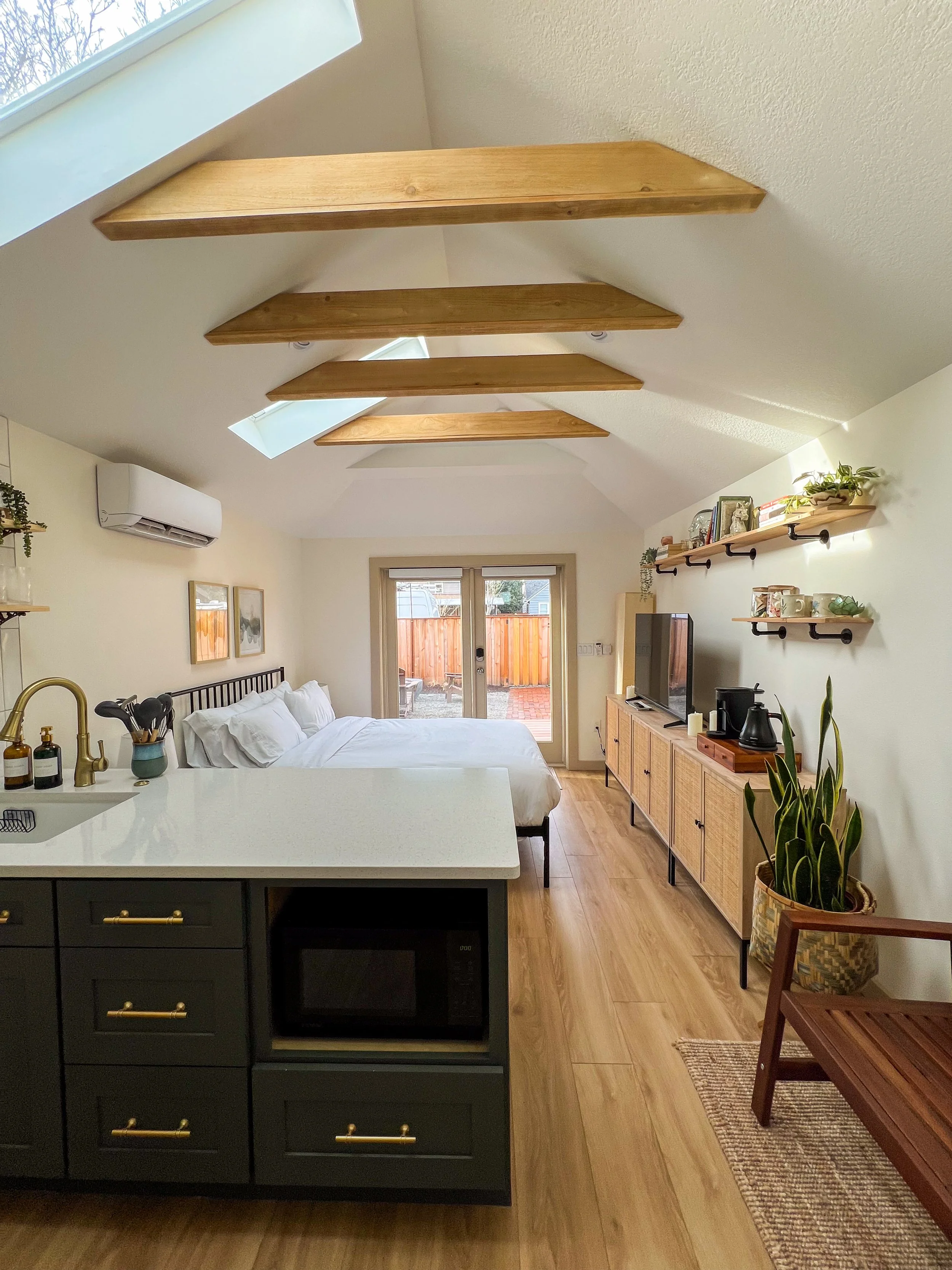 Interior view of a compact living space with a bed, kitchen counter, and a wooden sideboard. Features a sloped ceiling with exposed wooden beams and skylights. French doors lead outside, and a small seating area with a plant and shelves with decorative items.