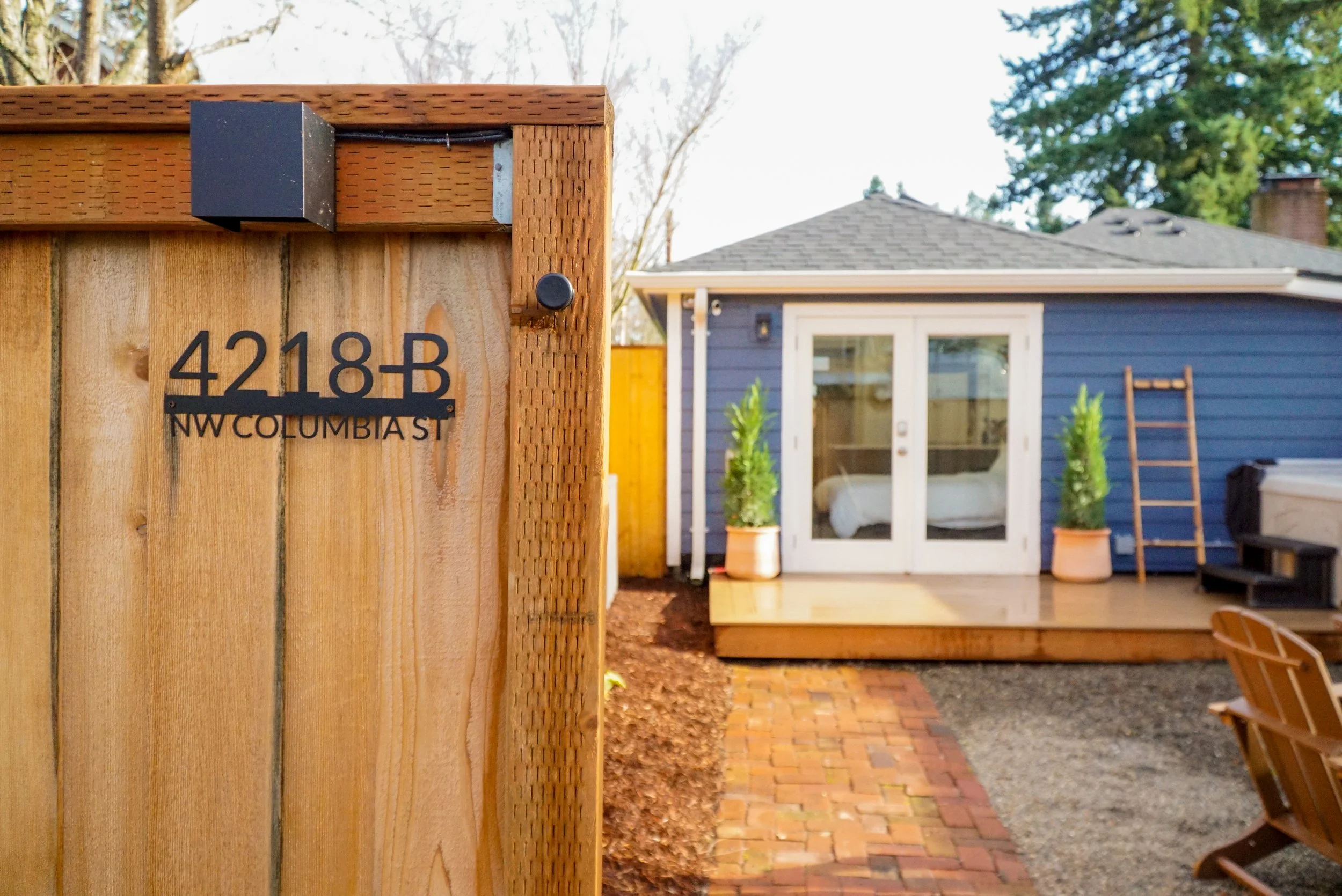 Close-up of a wooden fence with house number 4218-B and NW Columbia St sign, with a backyard deck, blue house, potted plants, and outdoor furniture in the background.