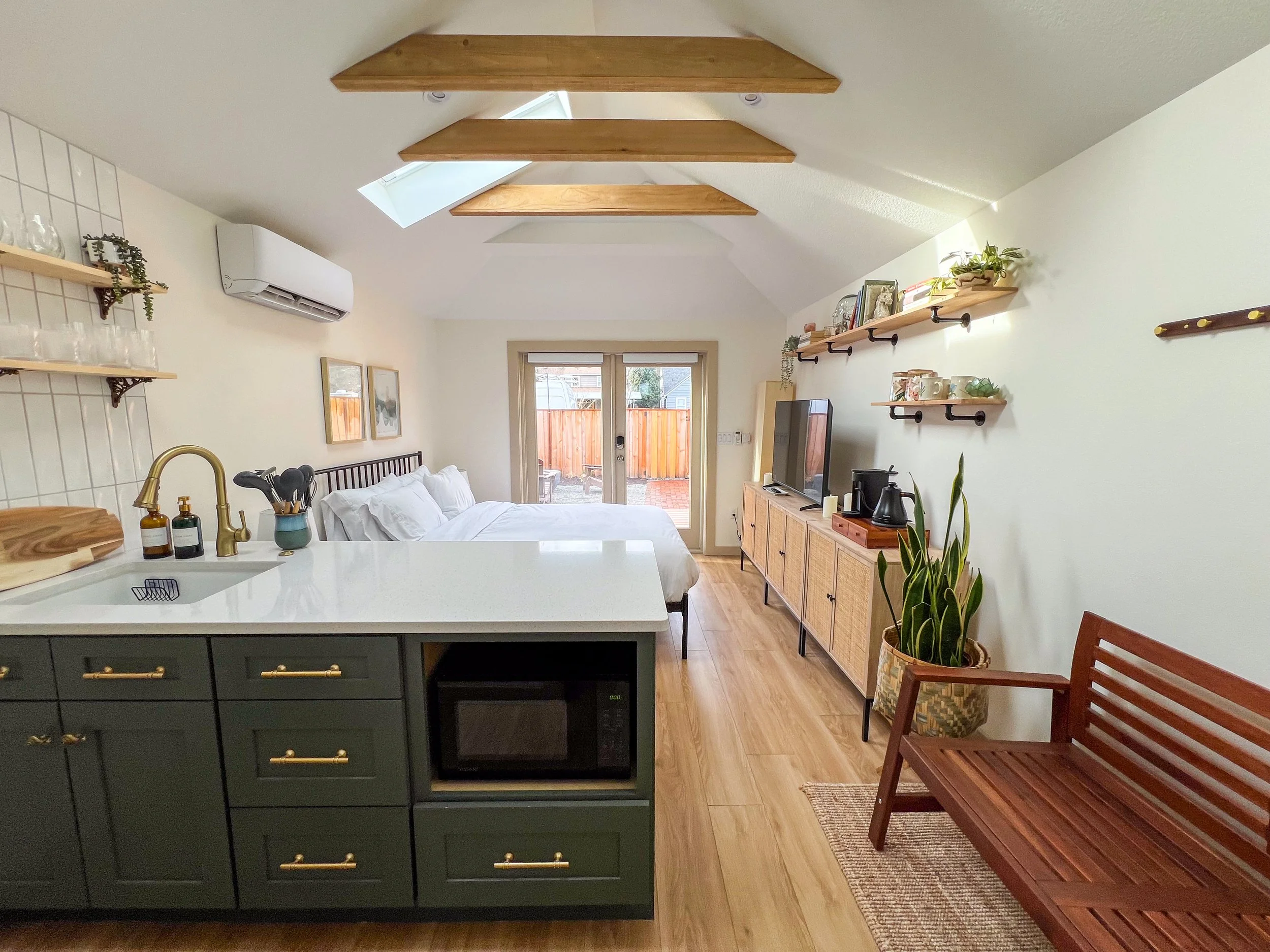 Open-concept studio apartment with a kitchen, bed, and living area. Kitchen features a green island with a microwave, sink, and gold handles. Bed with white linens is towards the back, with sliding glass doors leading to a backyard. Wooden shelves an