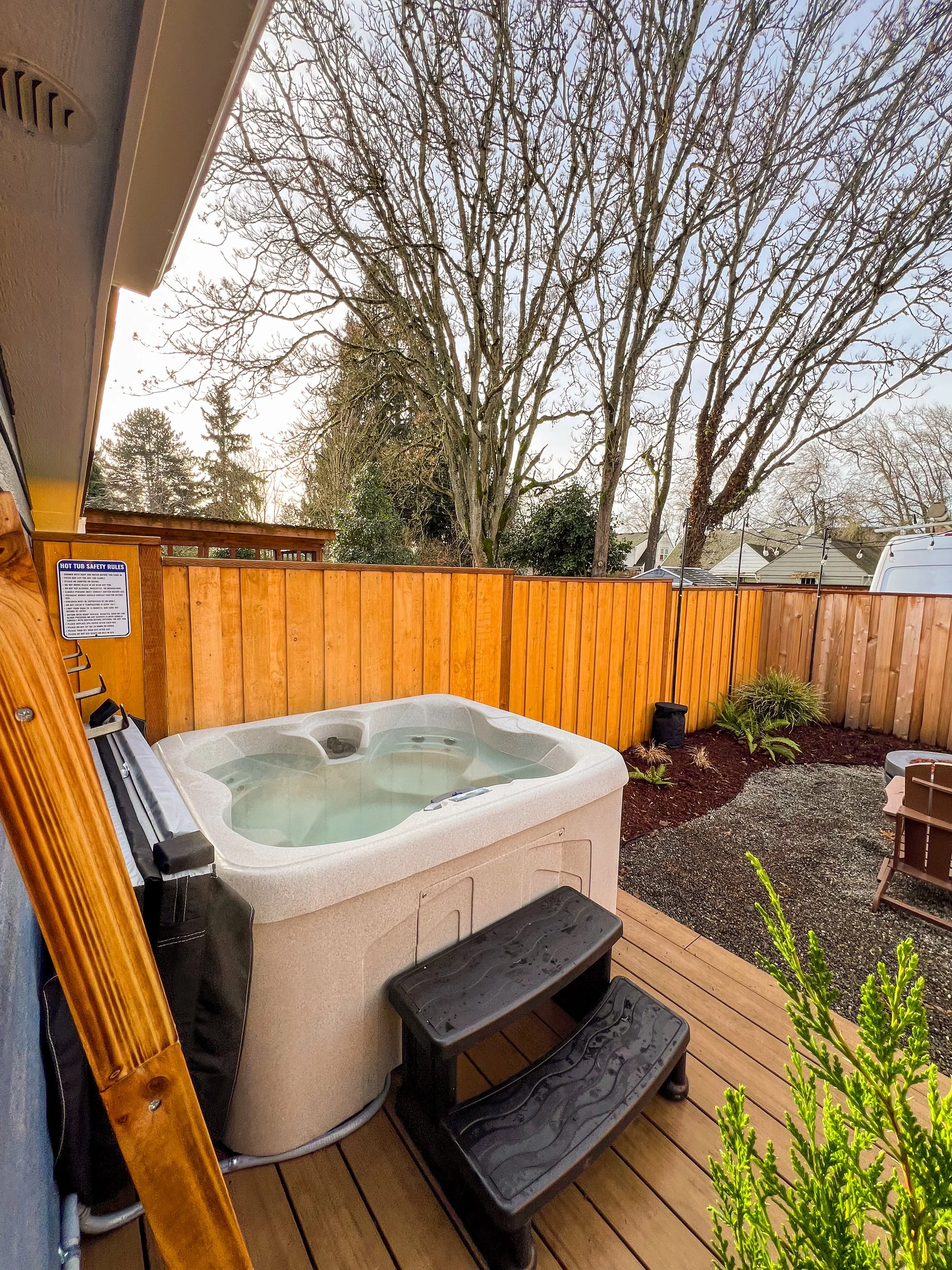 Backyard hot tub with wooden deck, surrounded by a wooden fence, trees without leaves, and outdoor furniture.