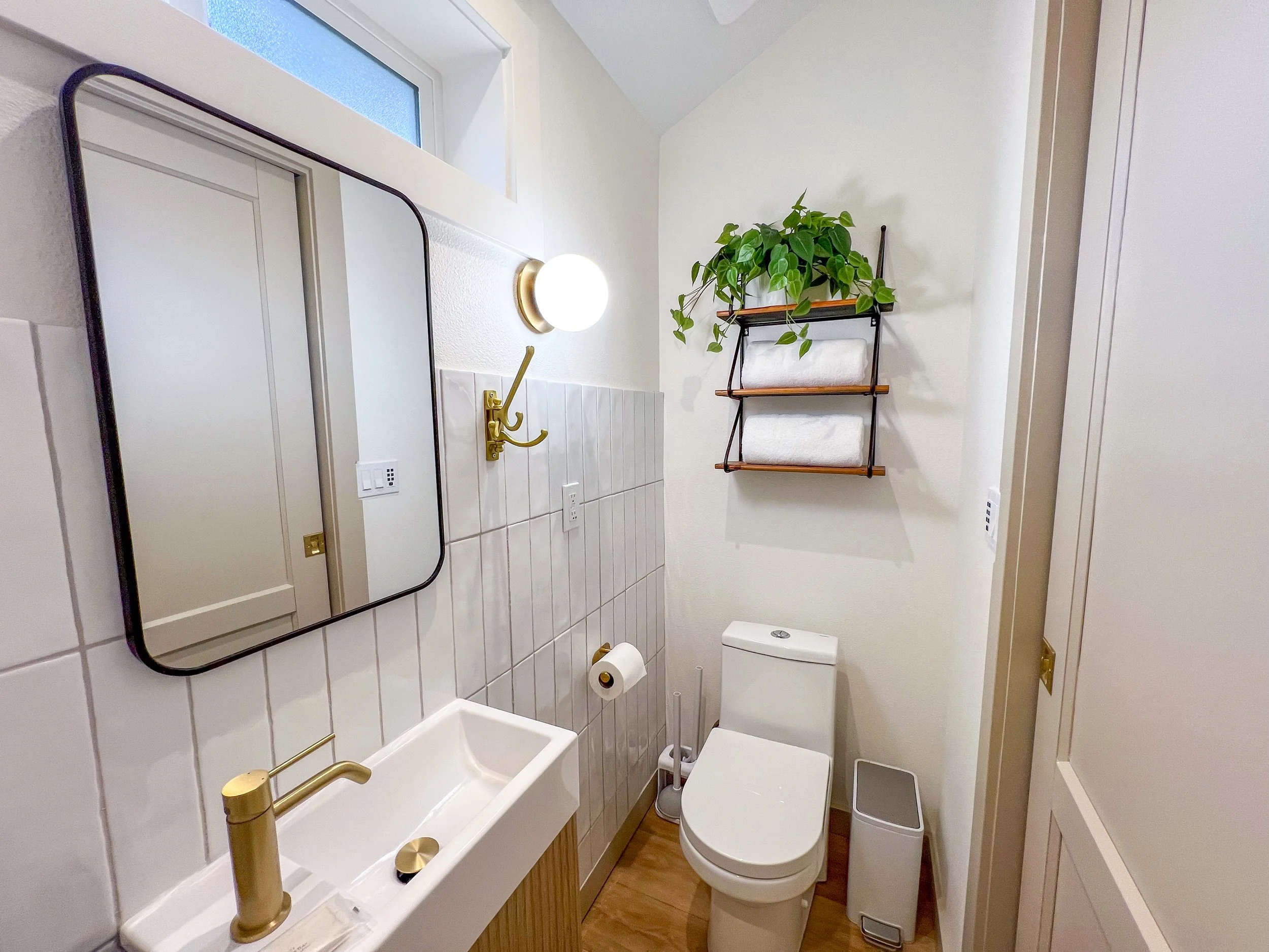 Small bathroom with white tiled sink, gold faucet, mirror, wall-mounted light, toilet, wall-mounted shelf with green plant and towels, small window, and a wooden floor.