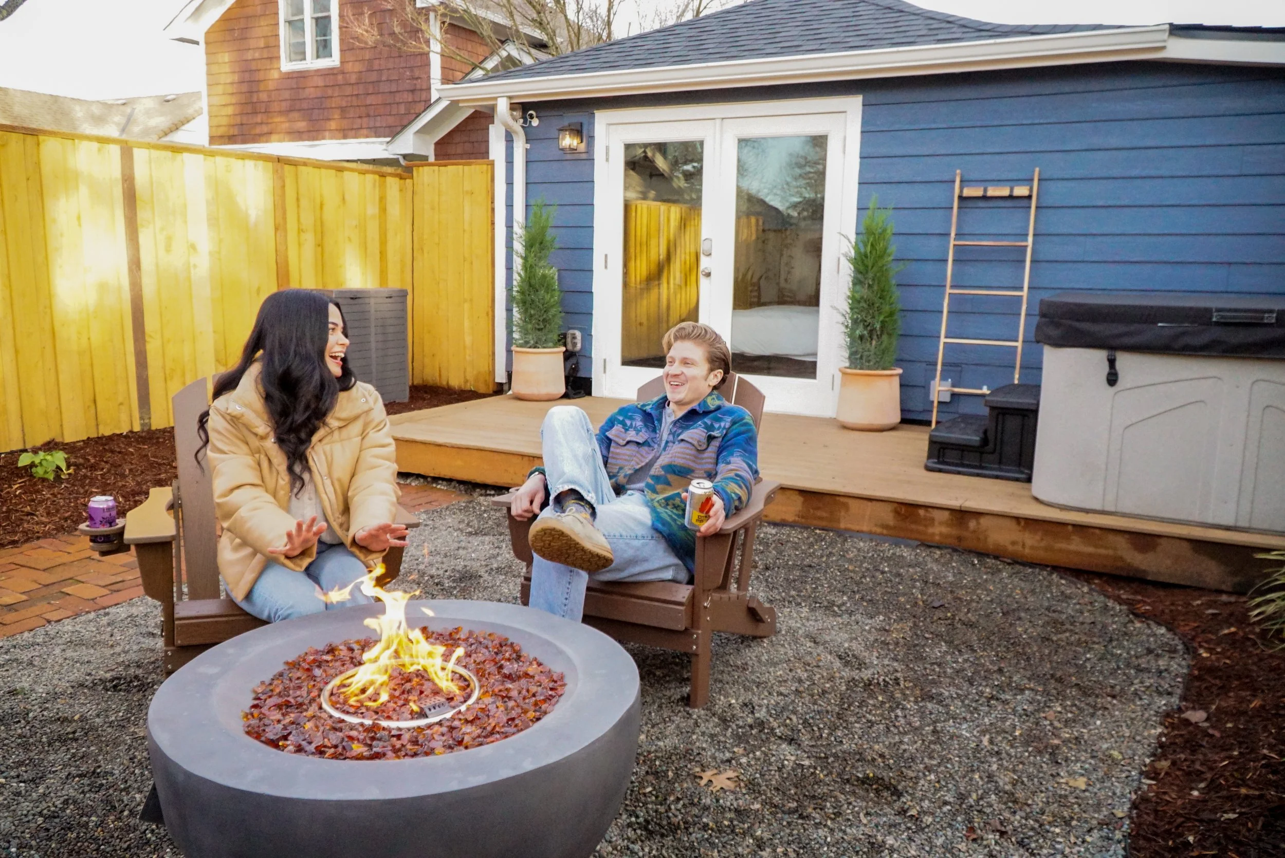 Two people sitting around a fire pit in a backyard, laughing and talking. One person wears a tan jacket and the other a patterned sweater. There are two potted plants and a blue house with a small deck in the background.