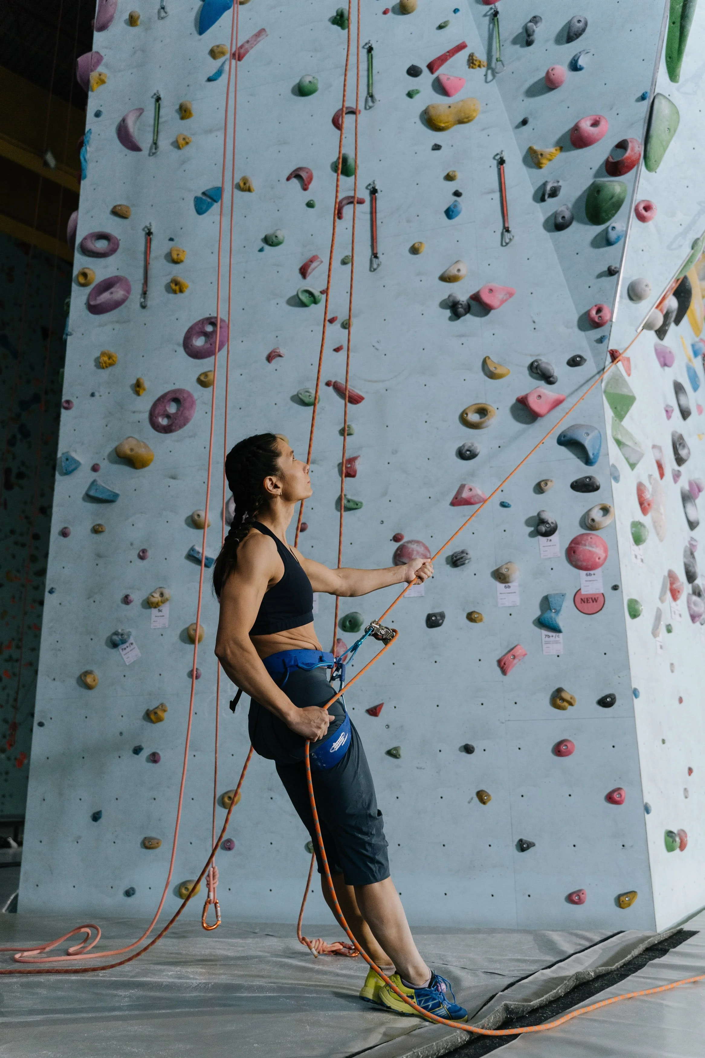A woman with a braided ponytail in a black sports top and black athletic pants using a harness to climb an indoor rock climbing wall with colorful holds.