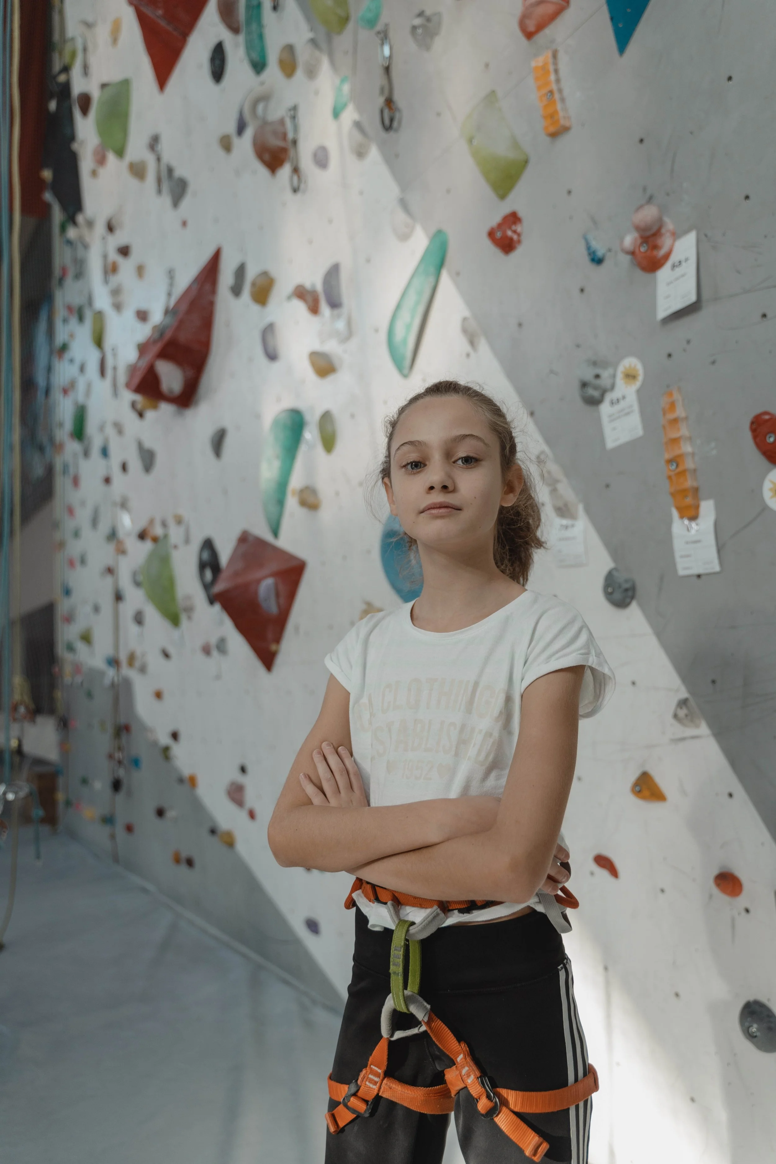 A young girl with crossed arms standing in front of an indoor rock climbing wall.