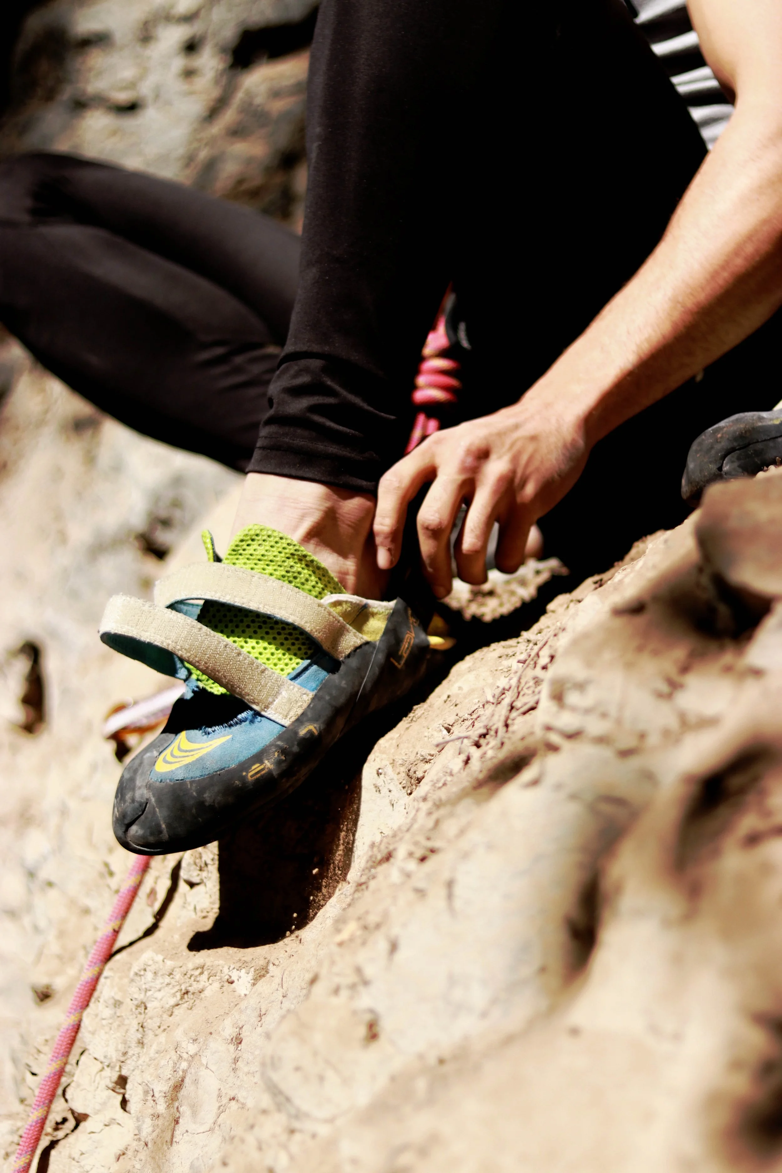 A person with a black and white striped shirt and black pants is sitting on rocky terrain, putting on or adjusting a climbing shoe with green, blue, and black colors, and beige velcro straps.