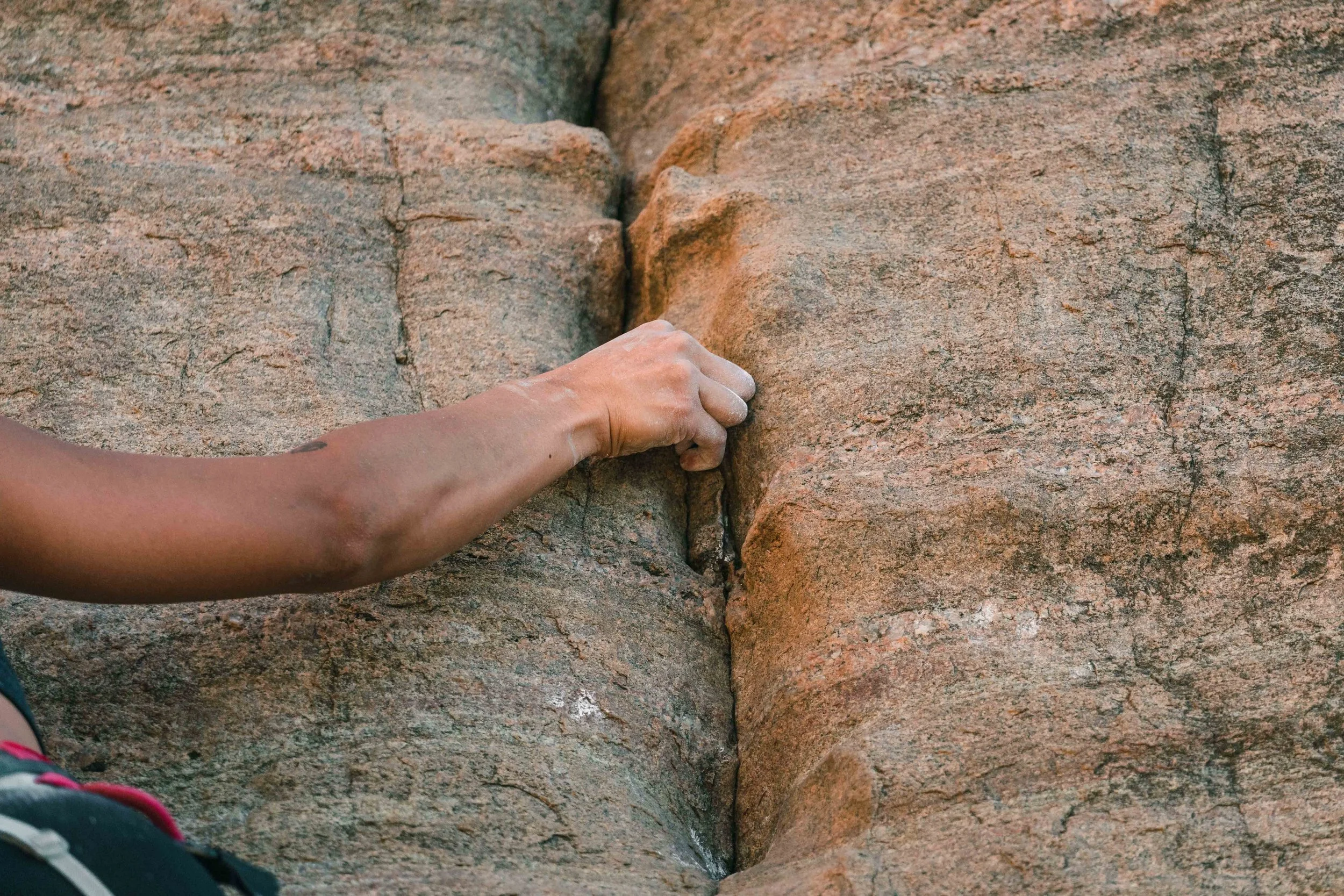 Close-up of a hand gripping a narrow crack in a reddish-brown rock formation during rock climbing.