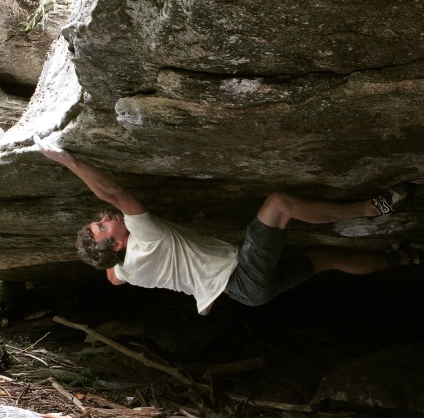 A man is bouldering on a large rock formation outdoors, wearing a white T-shirt, black shorts, and climbing shoes.