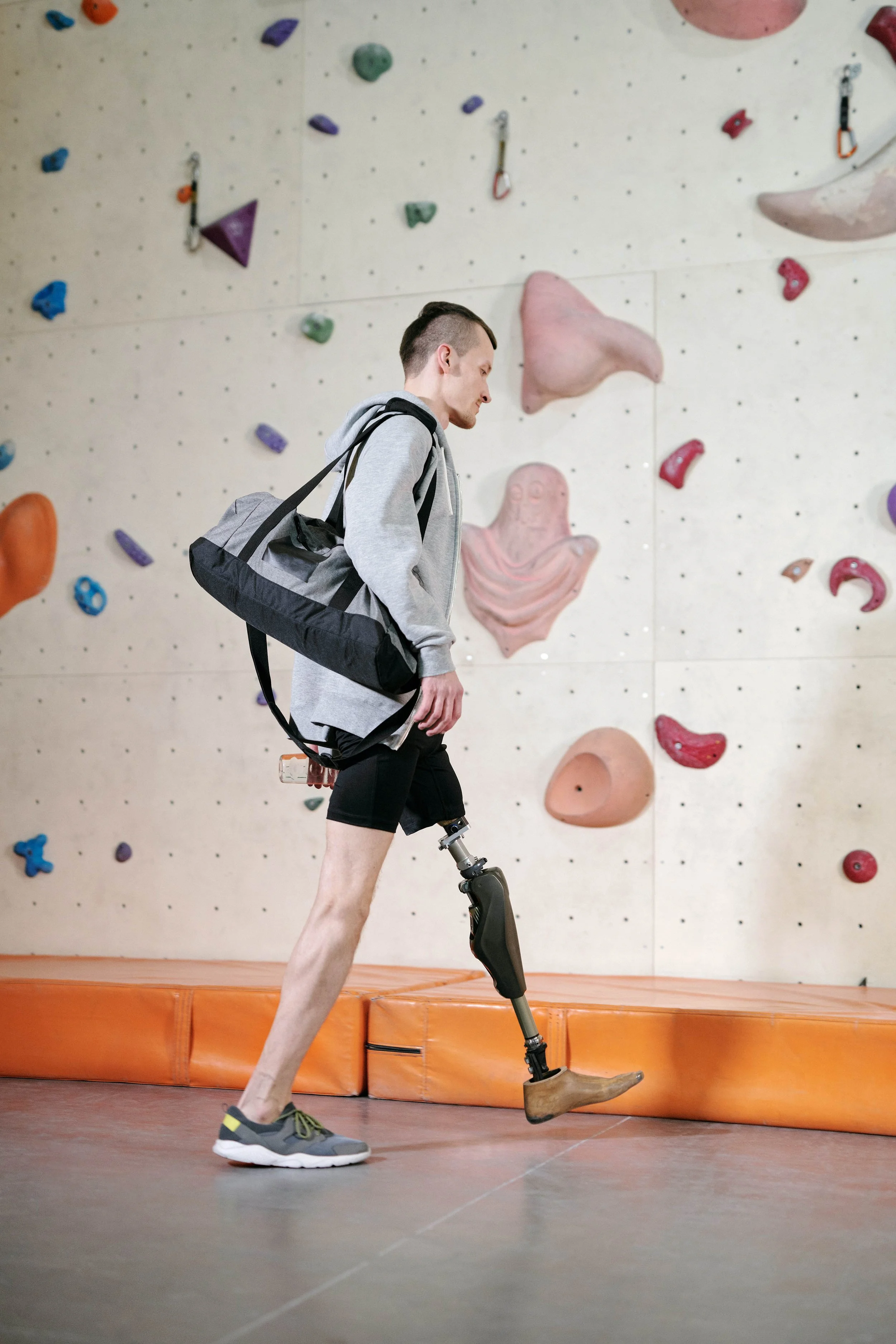 A man with a prosthetic leg walking inside a rock climbing gym with a climbing wall in the background.