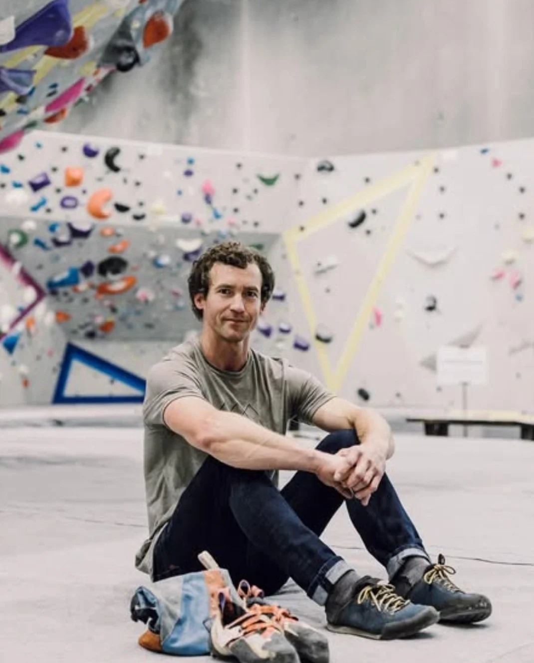 A man sitting on the floor of an indoor climbing gym with climbing shoes and a bag in front of him, with a rock climbing wall in the background.