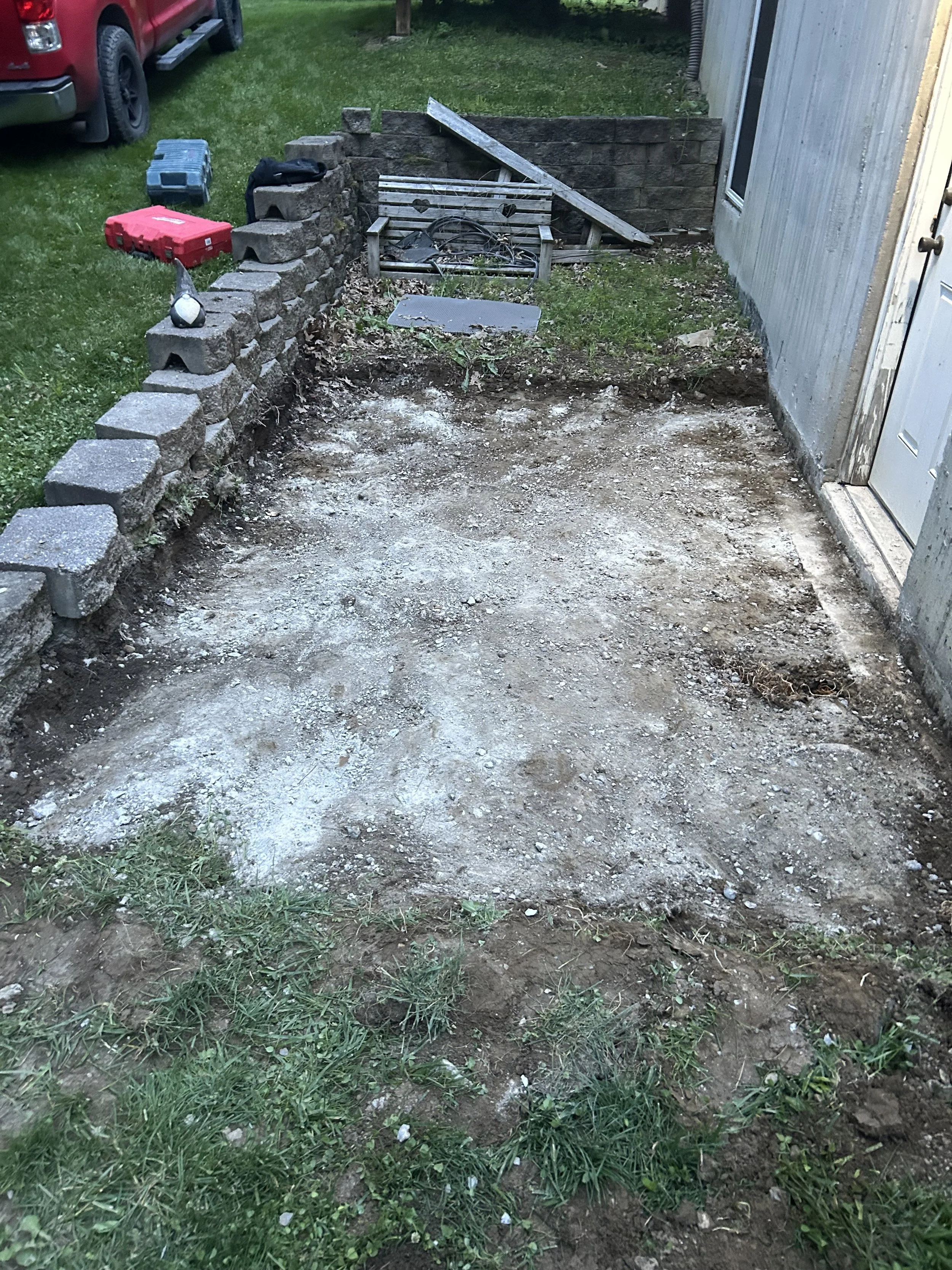A partially dug up yard with dirt and patches of grass, a small brick wall, a wooden bench, and a storage toolbox. A red truck is parked nearby, and a house is on the right side.