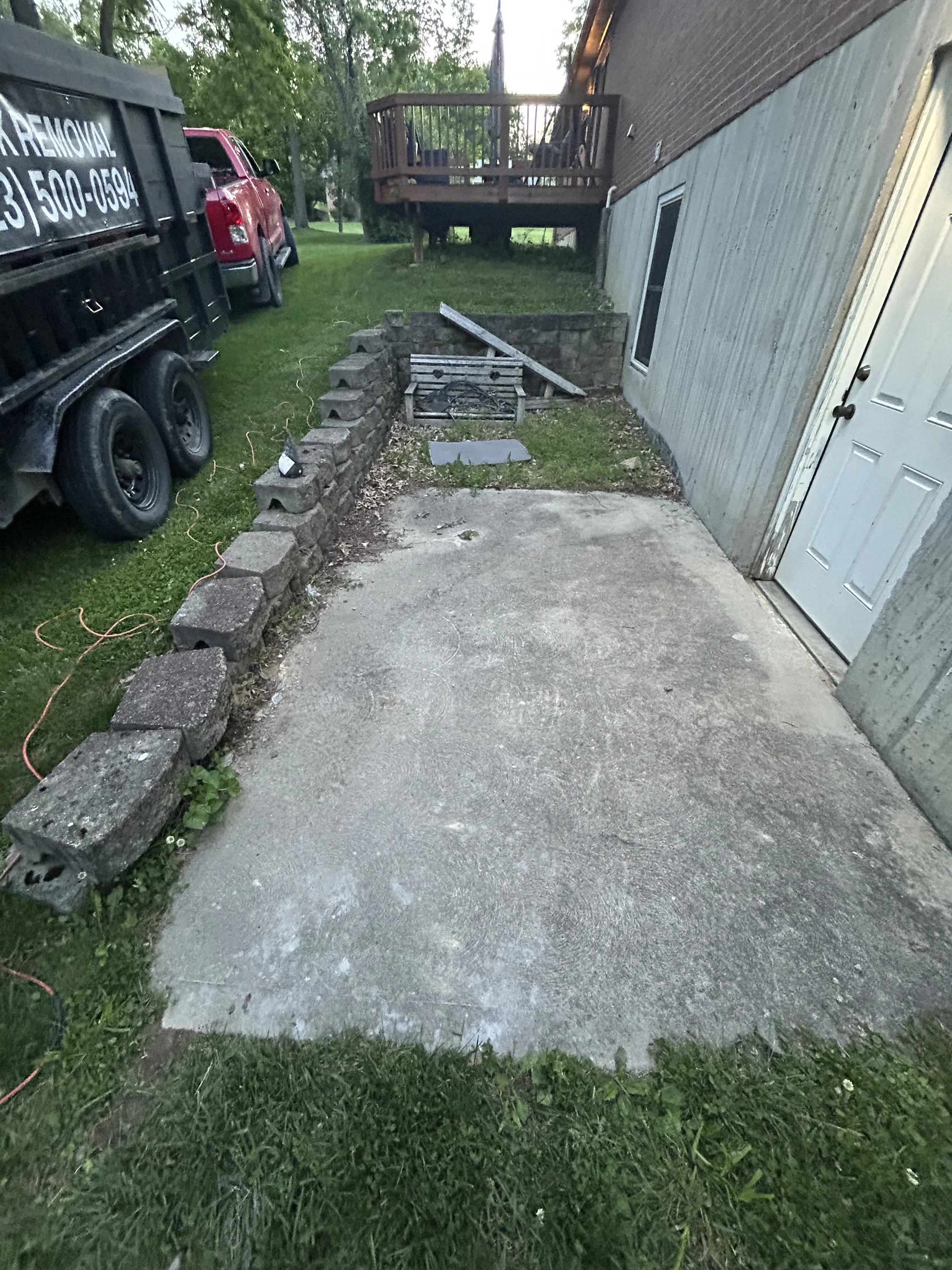 A small outdoor area with a grassy patch, surrounded by a brick and concrete wall, and a concrete sidewalk. To the left, there's a large black waste disposal truck and a red pickup truck. Behind, there's a wooden deck attached to a building with a door and a window. The area appears cluttered with some construction or yard work materials, including pieces of wood and a metal sheet.
