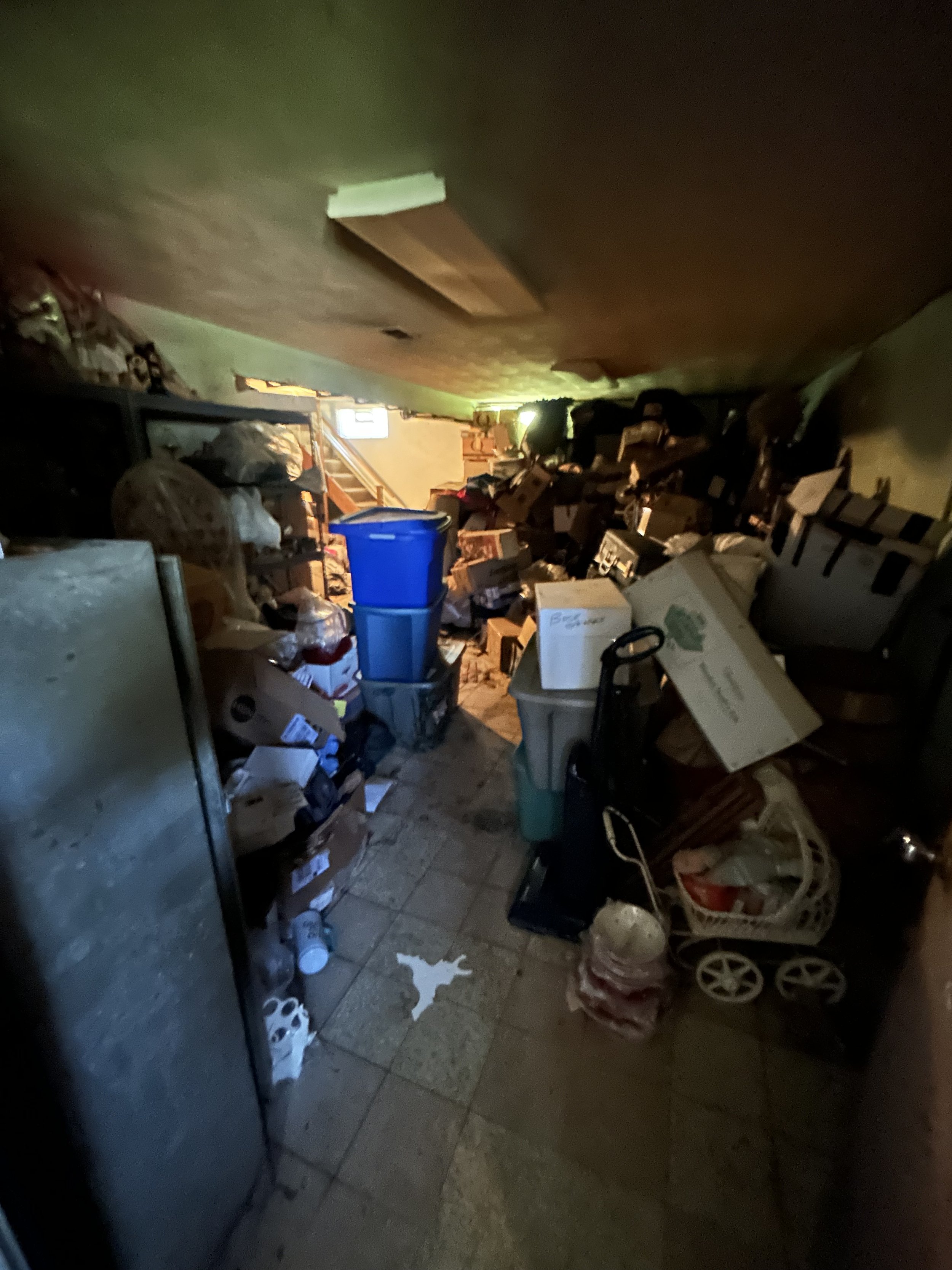 A cluttered storage room filled with boxes, bags, and miscellaneous household items, with a tiled floor and a small window.
