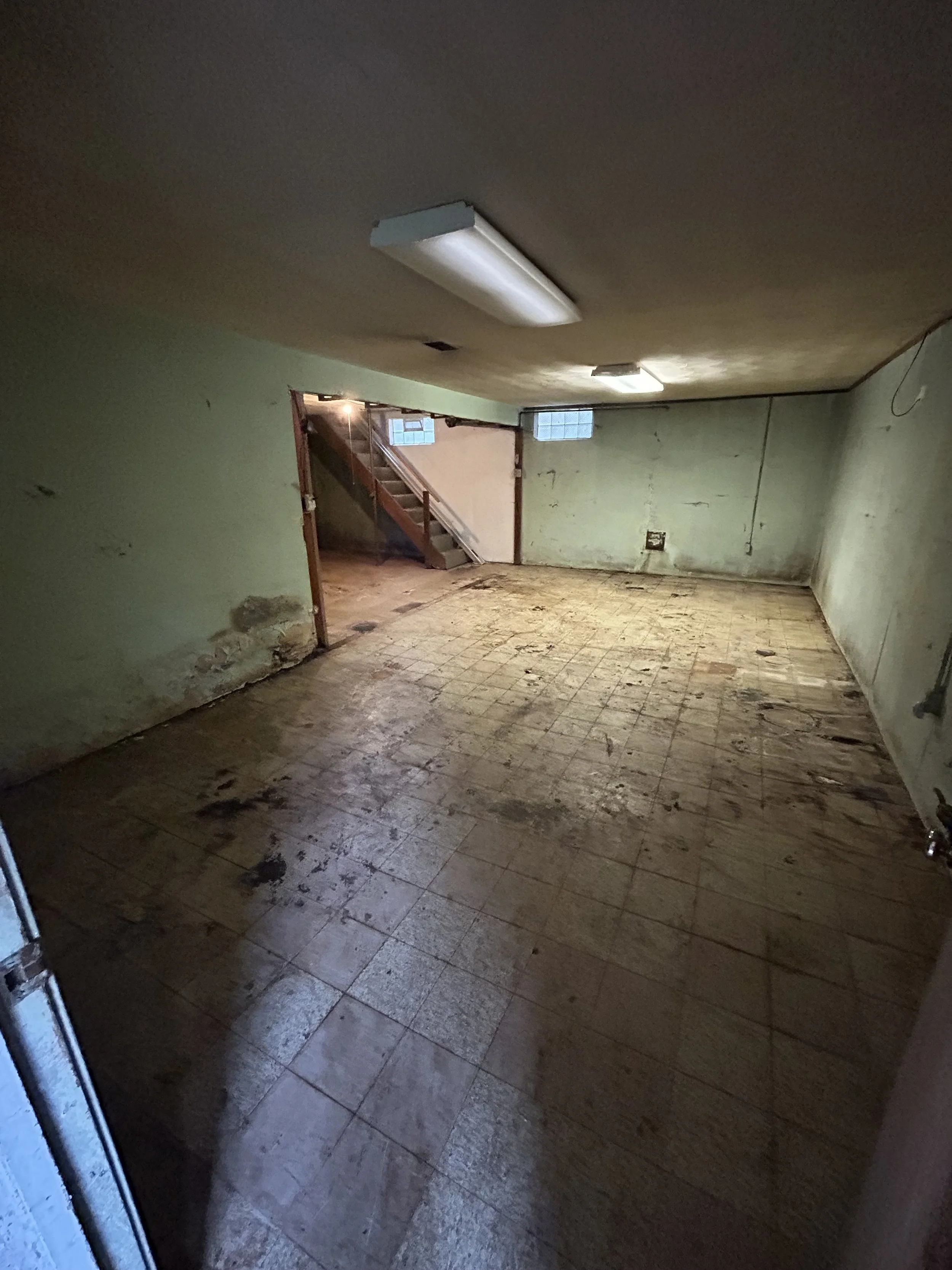 Empty room with greenish walls, a damaged floor, exposed electrical box, and ceiling lights, showing signs of neglect and disrepair.