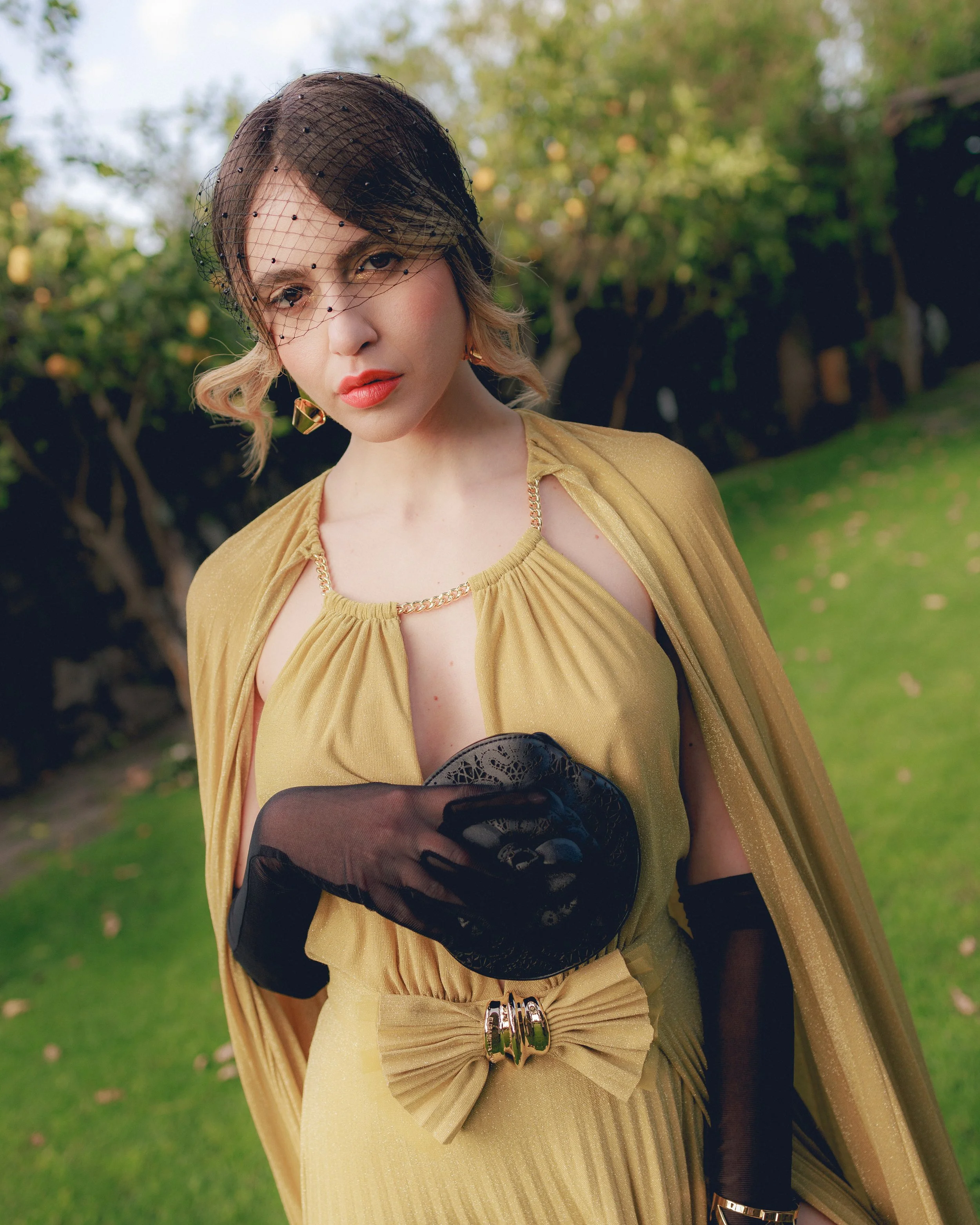 Una persona con un abito dorato elegante e accessori neri, inclusi guanti e un cappellino velato, in un giardino.