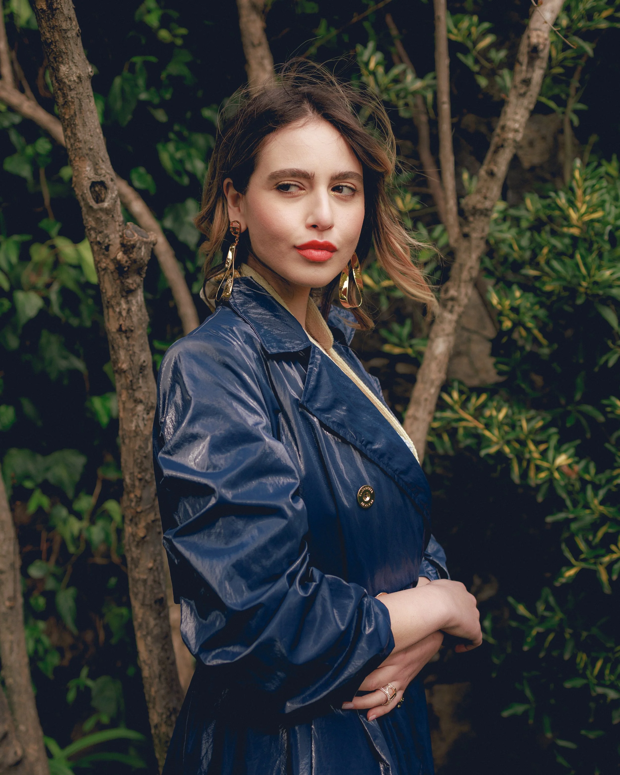Donna con cappotto blu e orecchini dorati, in piedi in un giardino con sfondo di piante.