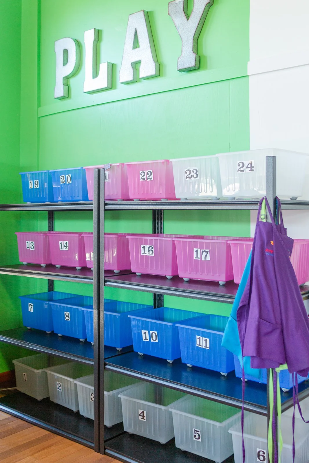 Colorful storage bins on a metal shelving unit, each labeled with numbers, in a playroom or daycare setting with a green wall and a large 'PLAY' sign.
