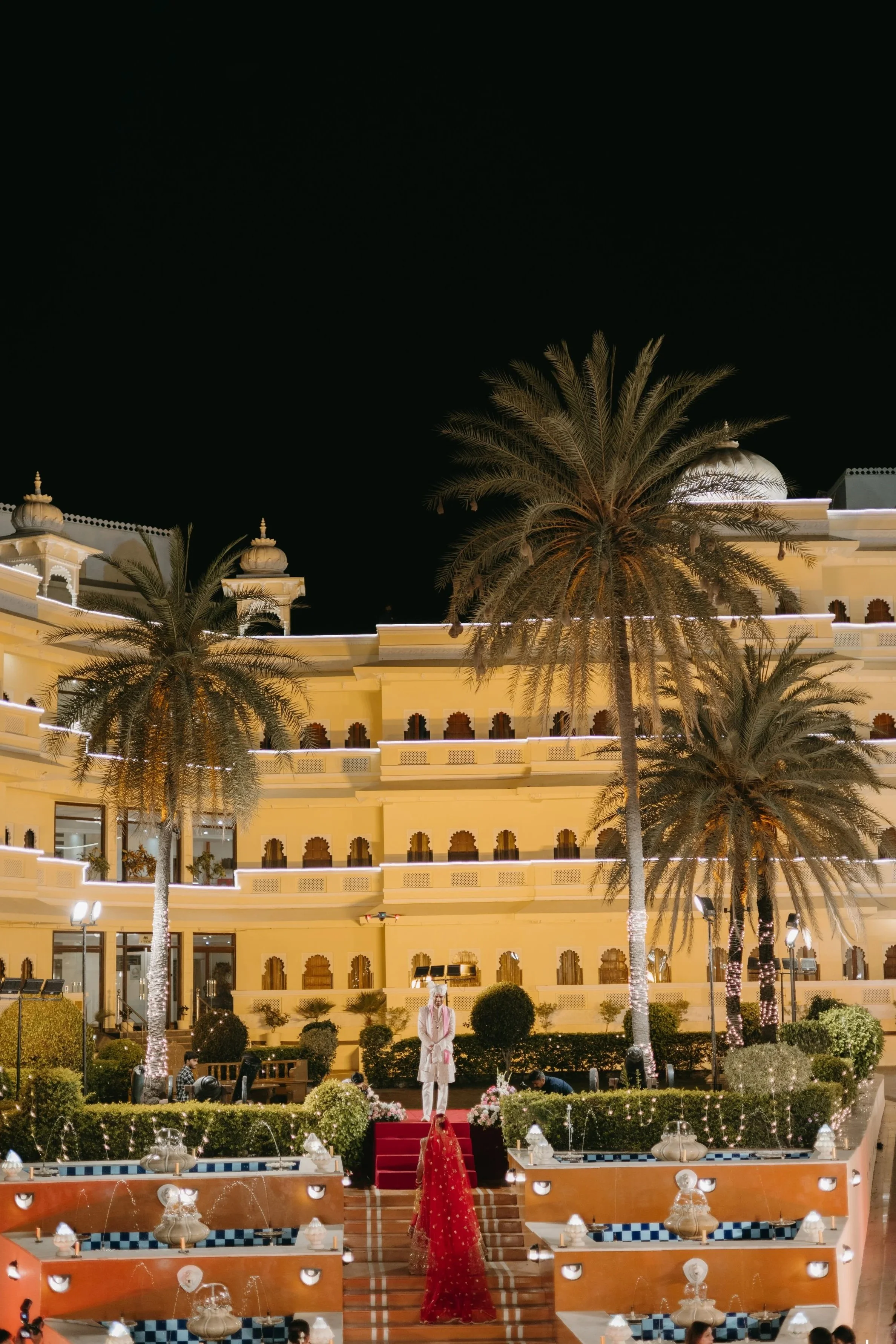 Nighttime view of a grand building with Indian-style architectural features, illuminated with string lights and surrounded by palm trees. In the foreground, a stage with steps, decorated with lights and water fountains, features a person dressed in traditional Indian attire standing on a red carpet.
