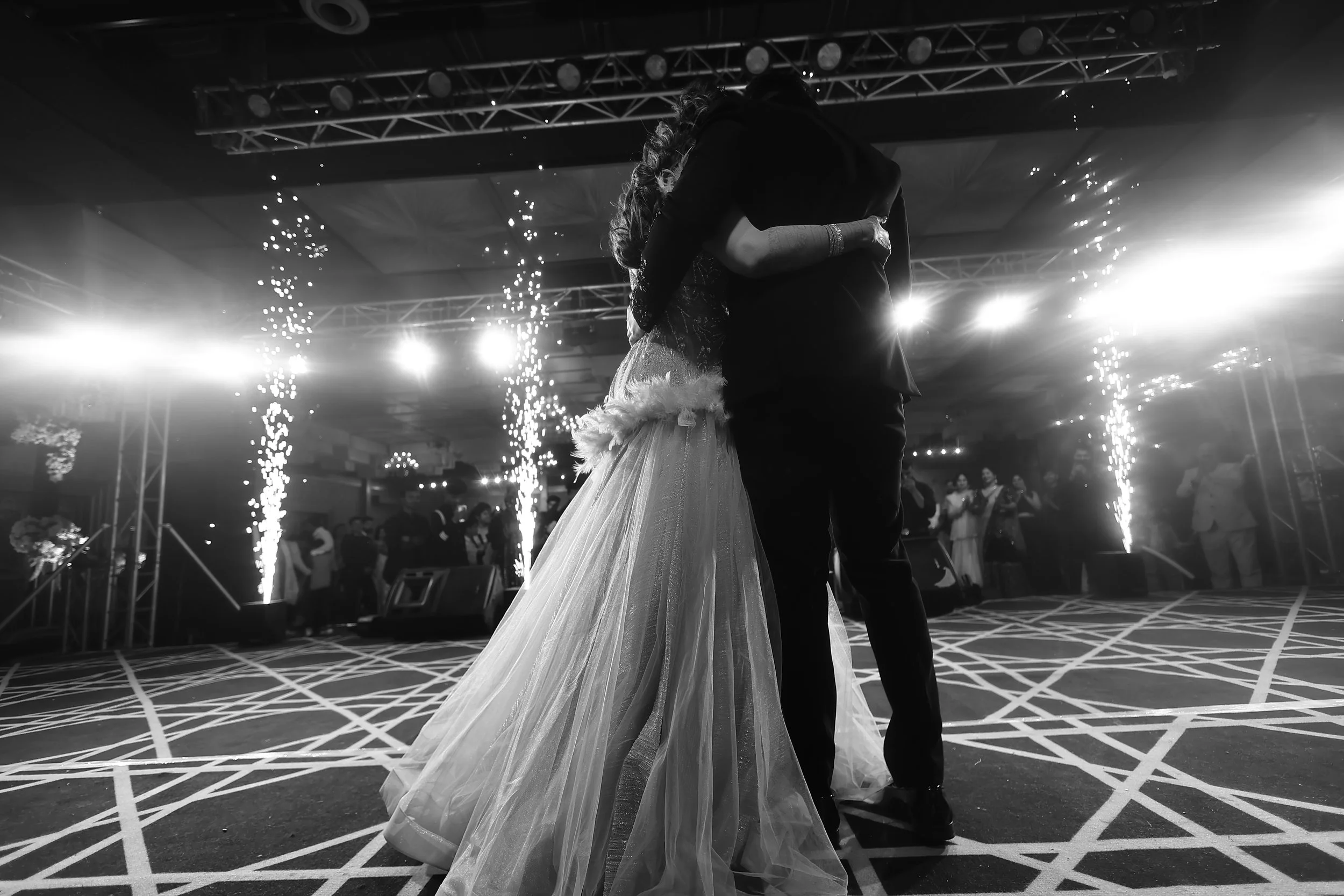 A bride and groom dancing at their wedding reception, embraced closely. The scene is illuminated by bright lights and fireworks, with guests watching in the background.