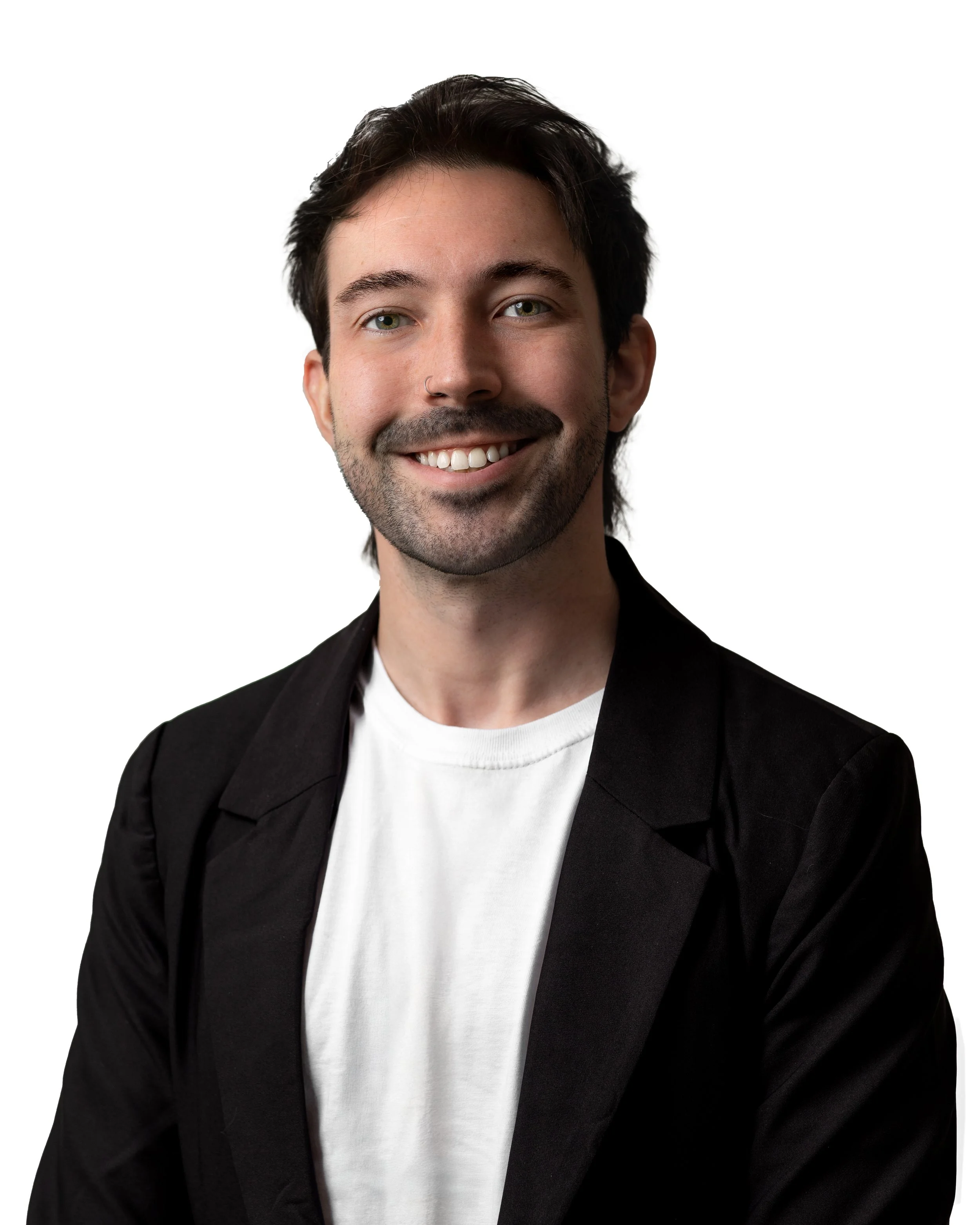 A smiling man with dark hair, a beard, wearing a black blazer and white t-shirt, against a white background.