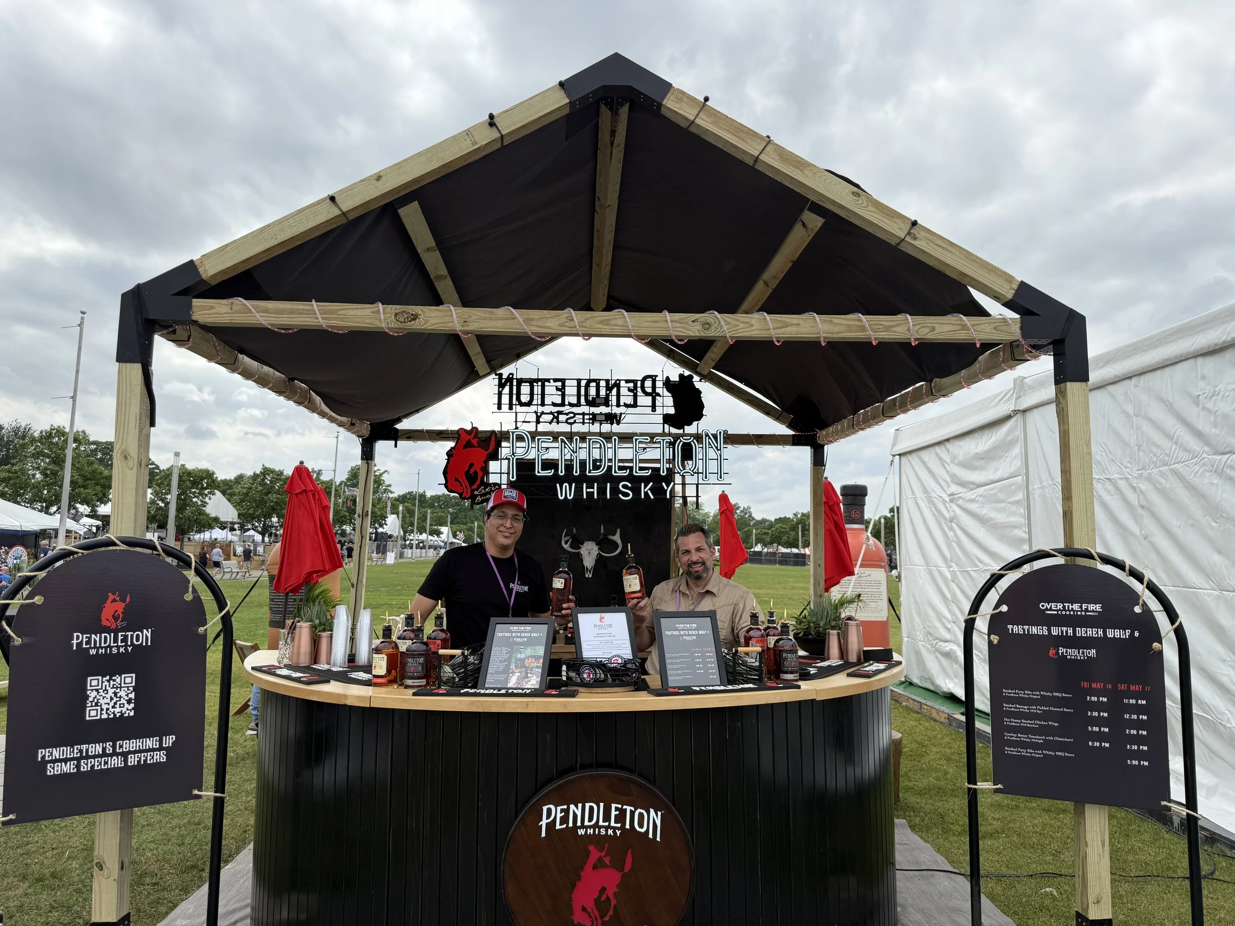 At an outdoor event, a whiskey tasting booth is set up under a wooden and fabric canopy. Two men are standing behind the counter, smiling and holding whiskey bottles. The booth has signage for Pendleton Whisky, featuring a red horse logo and a neon sign. There are menus, branded cups, and whiskey bottles on the counter. The background shows a grassy field with tents and trees under a cloudy sky.