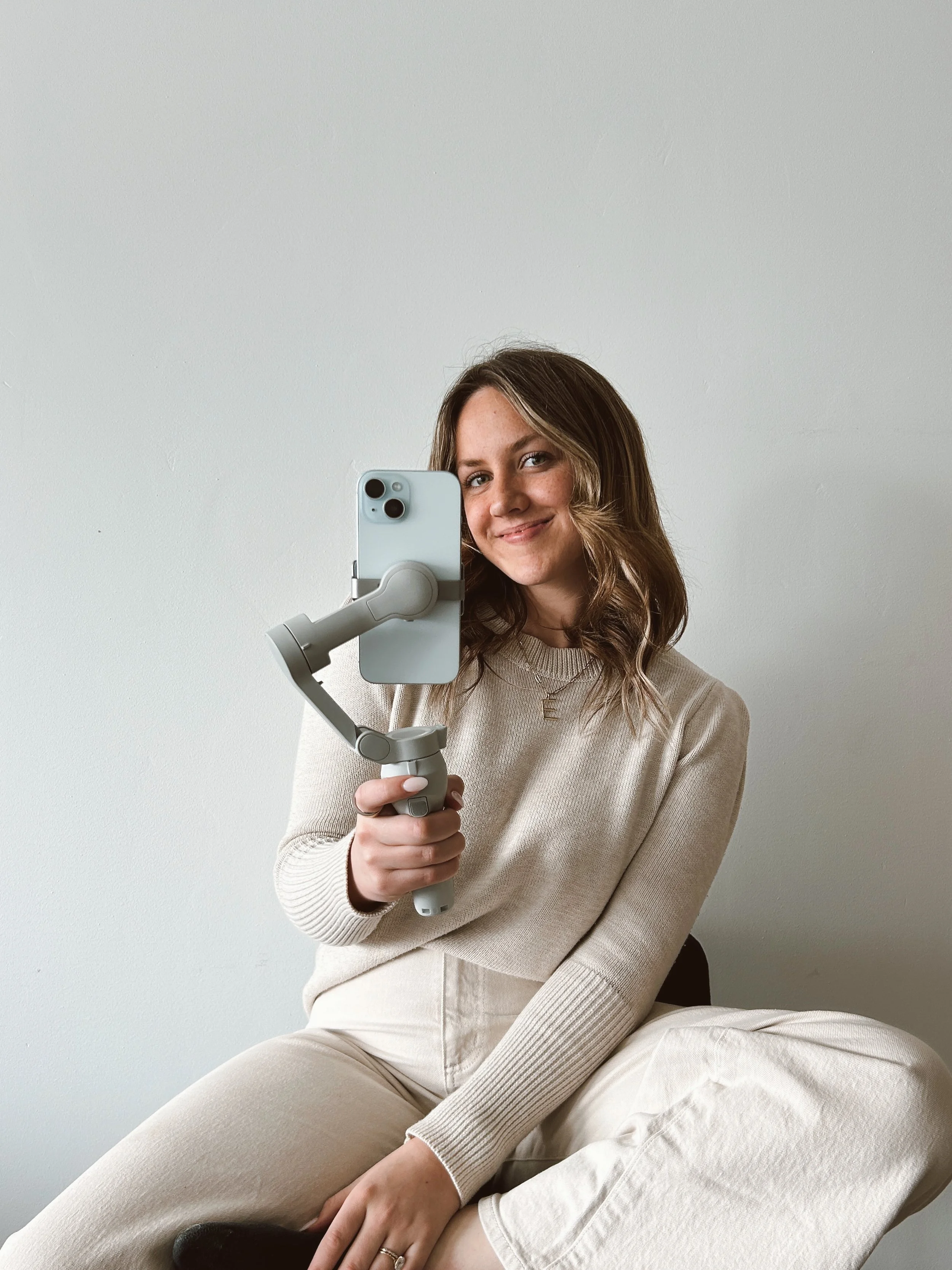 Person taking a selfie with a smartphone on a gimbal, wearing a beige outfit, against a plain background.