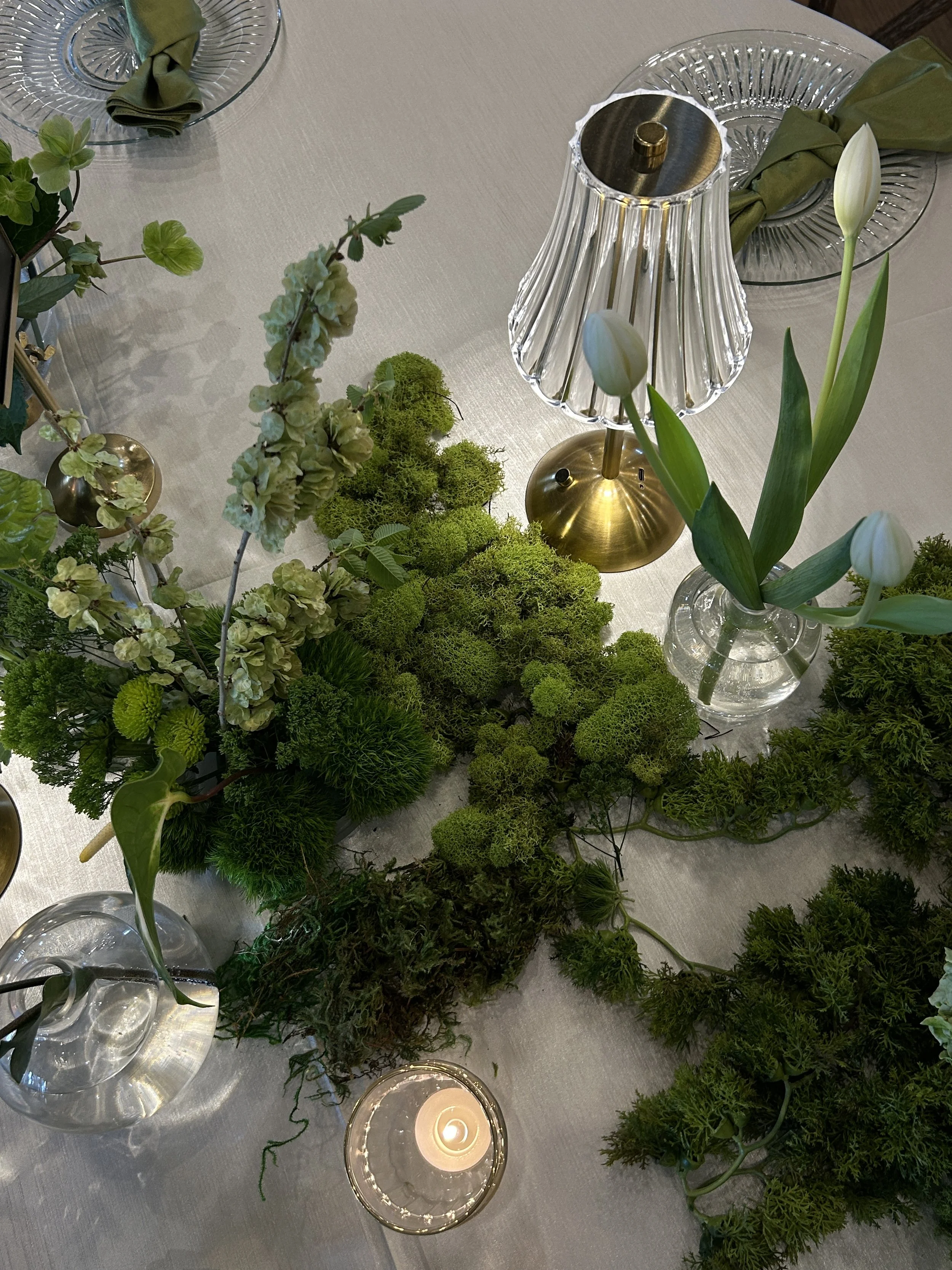 Elegant banquet hall with long tables decorated with white tablecloths, floral centerpieces, and candles. Black chairs are arranged around the tables, and windows provide natural light. Hanging flowers adorn the ceiling.