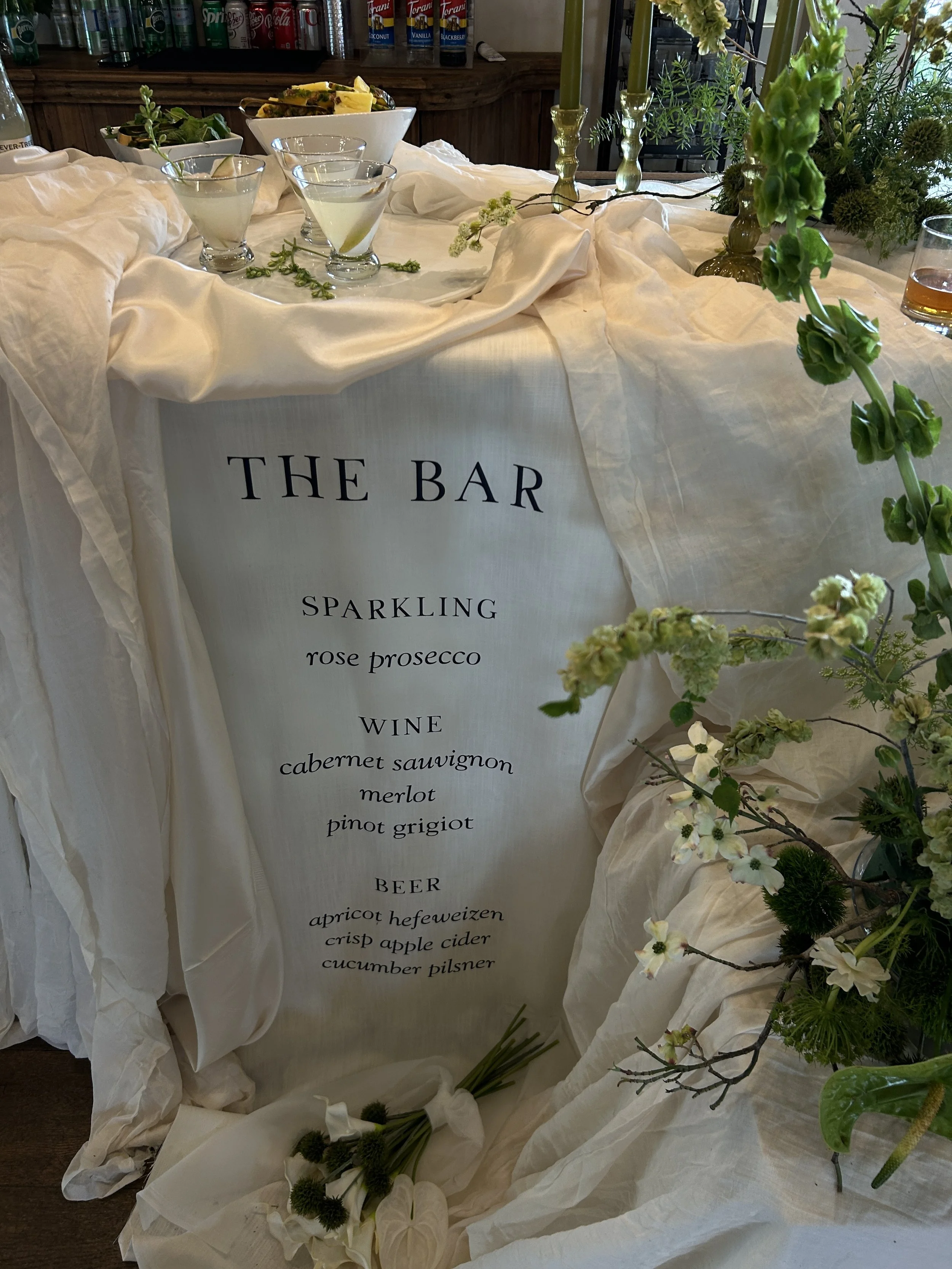 Outdoor wedding venue with sign reading "Welcome to the wedding of Alexis and Brandon" on an easel, surrounded by trees and a building in the background.