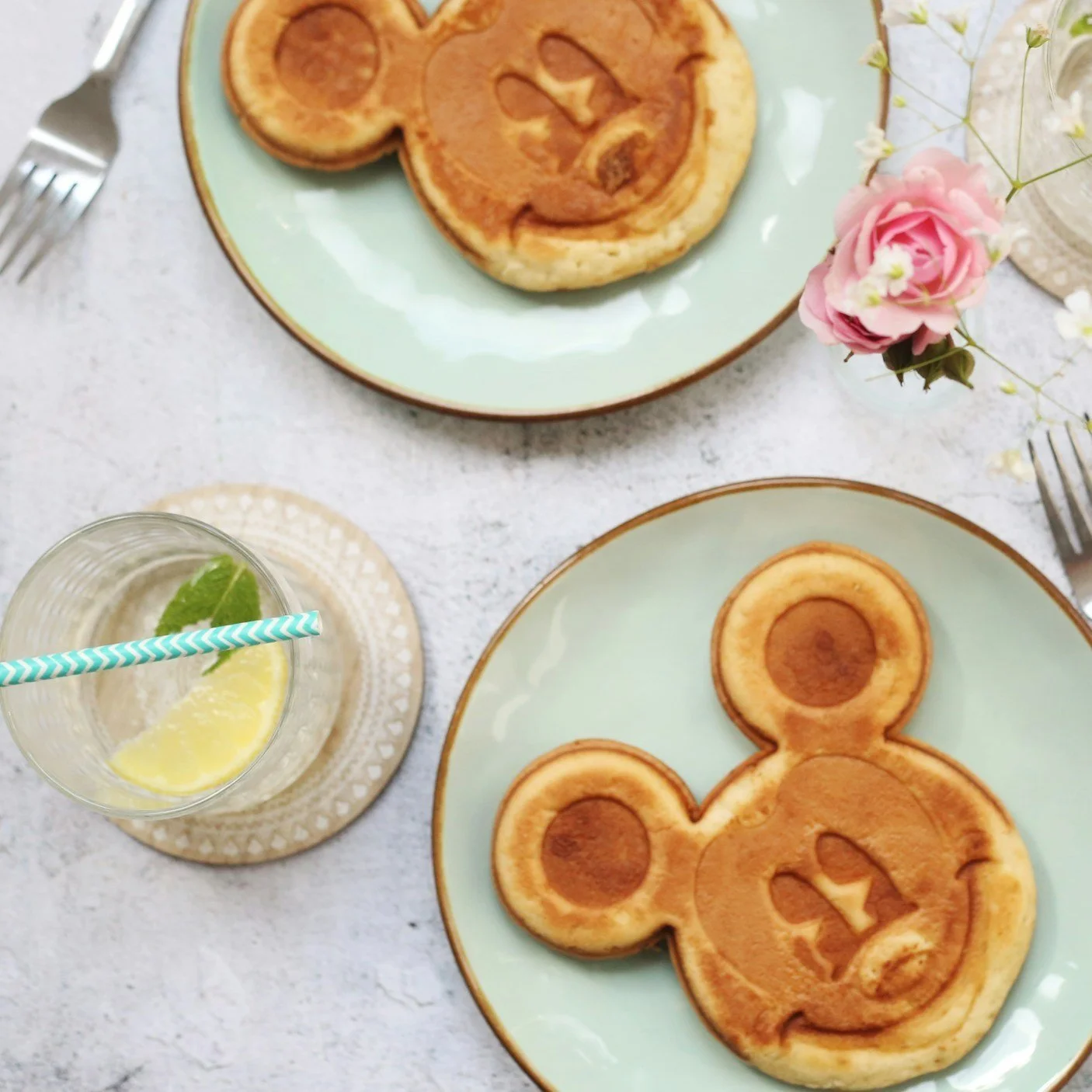 Cartoon character-shaped pancakes on green plates with flowers and a lemon mint drink on a gray surface.