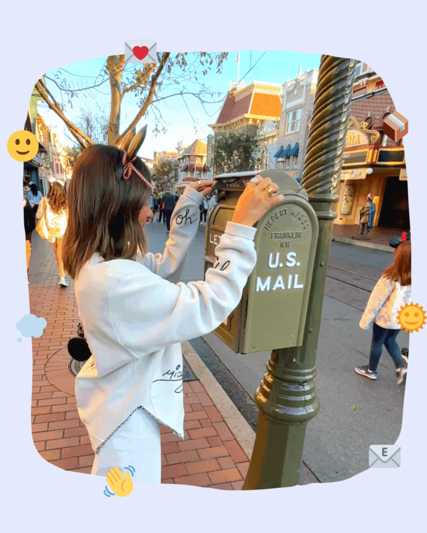 Person mailing a letter in a vintage U.S. mailbox on a street with other people walking by, decorated with emojis and stickers.