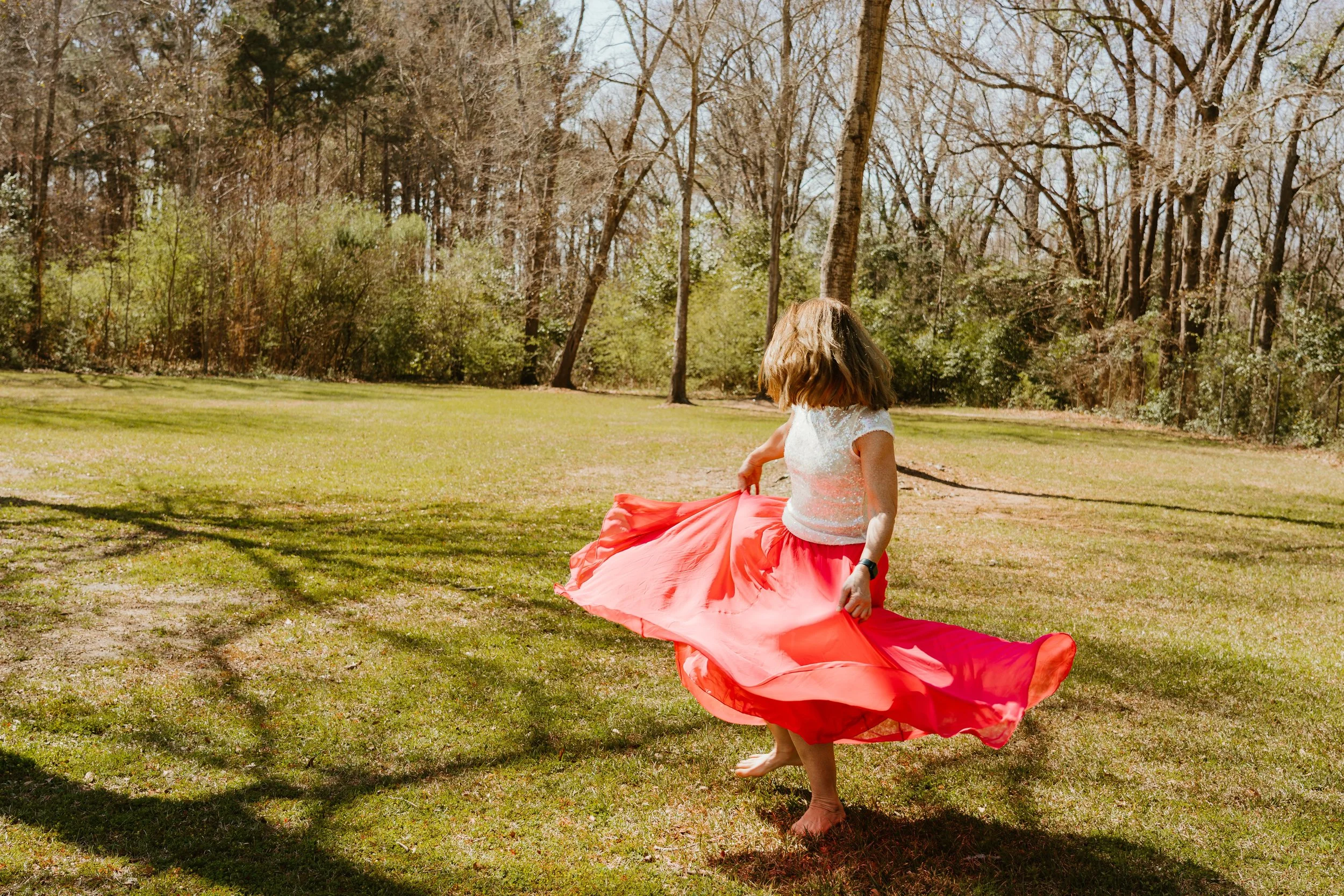 A young girl twirling in a grassy park on a sunny day, wearing a white top and a long, flowing coral skirt.