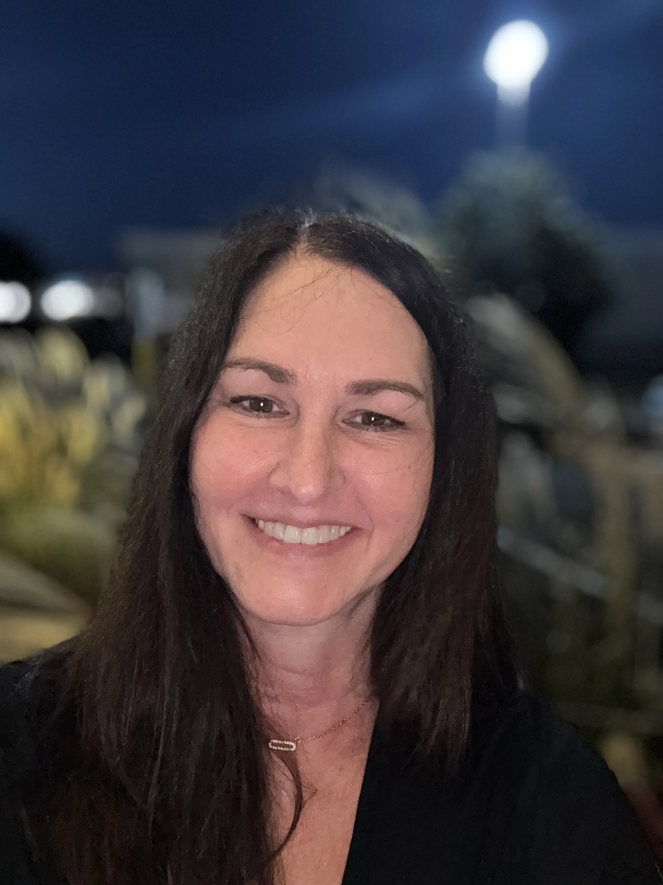 A smiling woman with long dark hair standing outside at night with a bright moon in the sky.