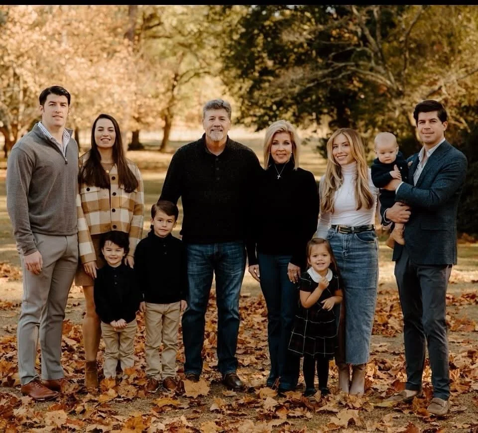 A family of ten posing outdoors in a park during fall, surrounded by trees with autumn leaves on the ground, smiling and standing close together.