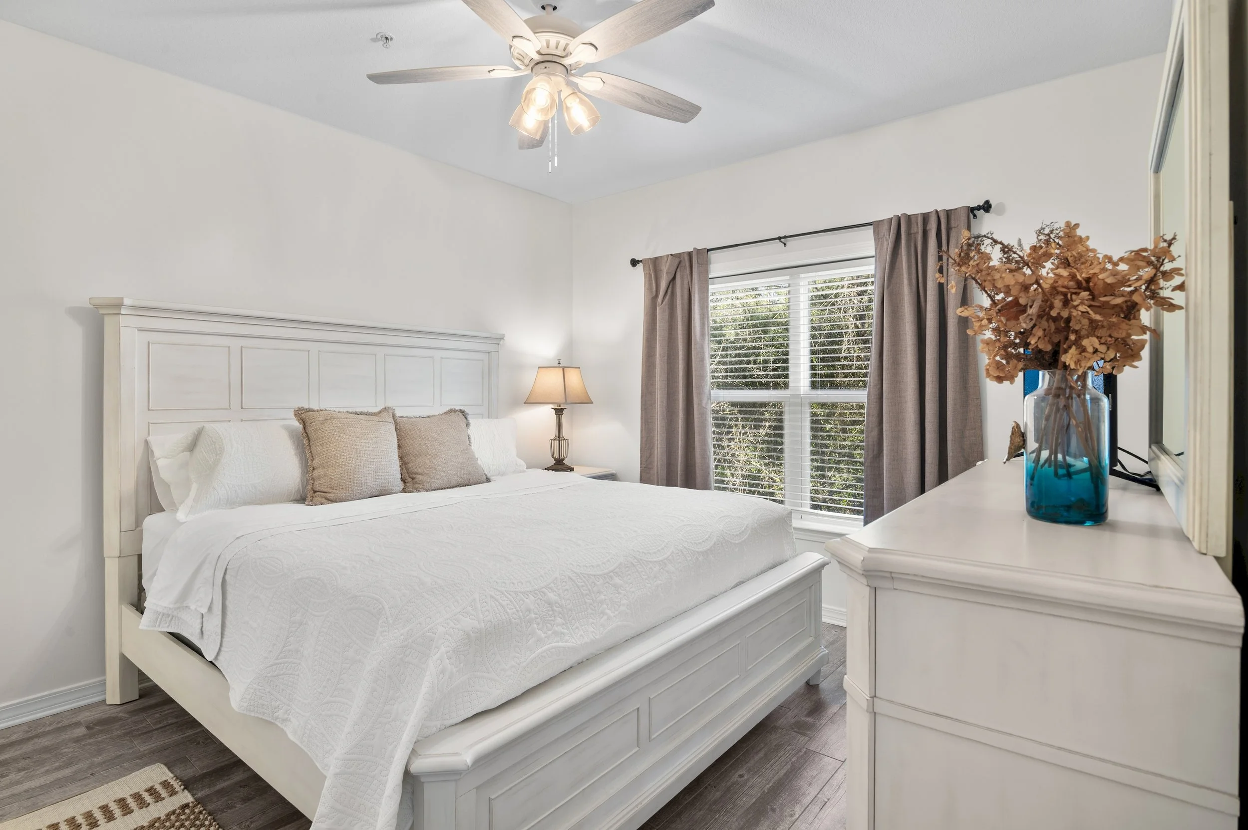 Bedroom with white bed, beige pillows, wooden nightstand with lamp, window with blinds and curtains, dresser with blue vase and dried flowers, ceiling fan.