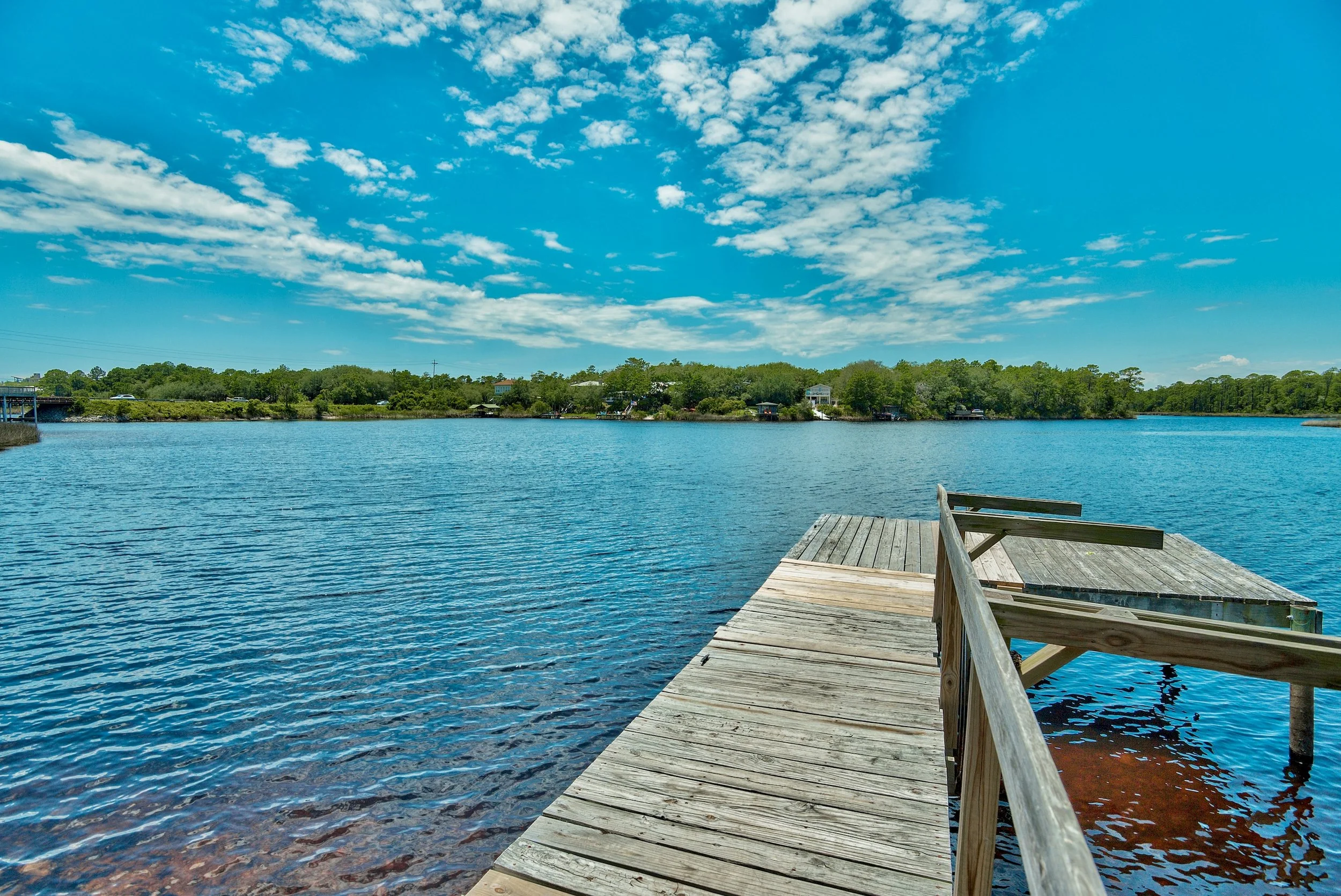 A wooden dock extends into a calm lake with blue water, under a partly cloudy sky, with trees and houses in the background.