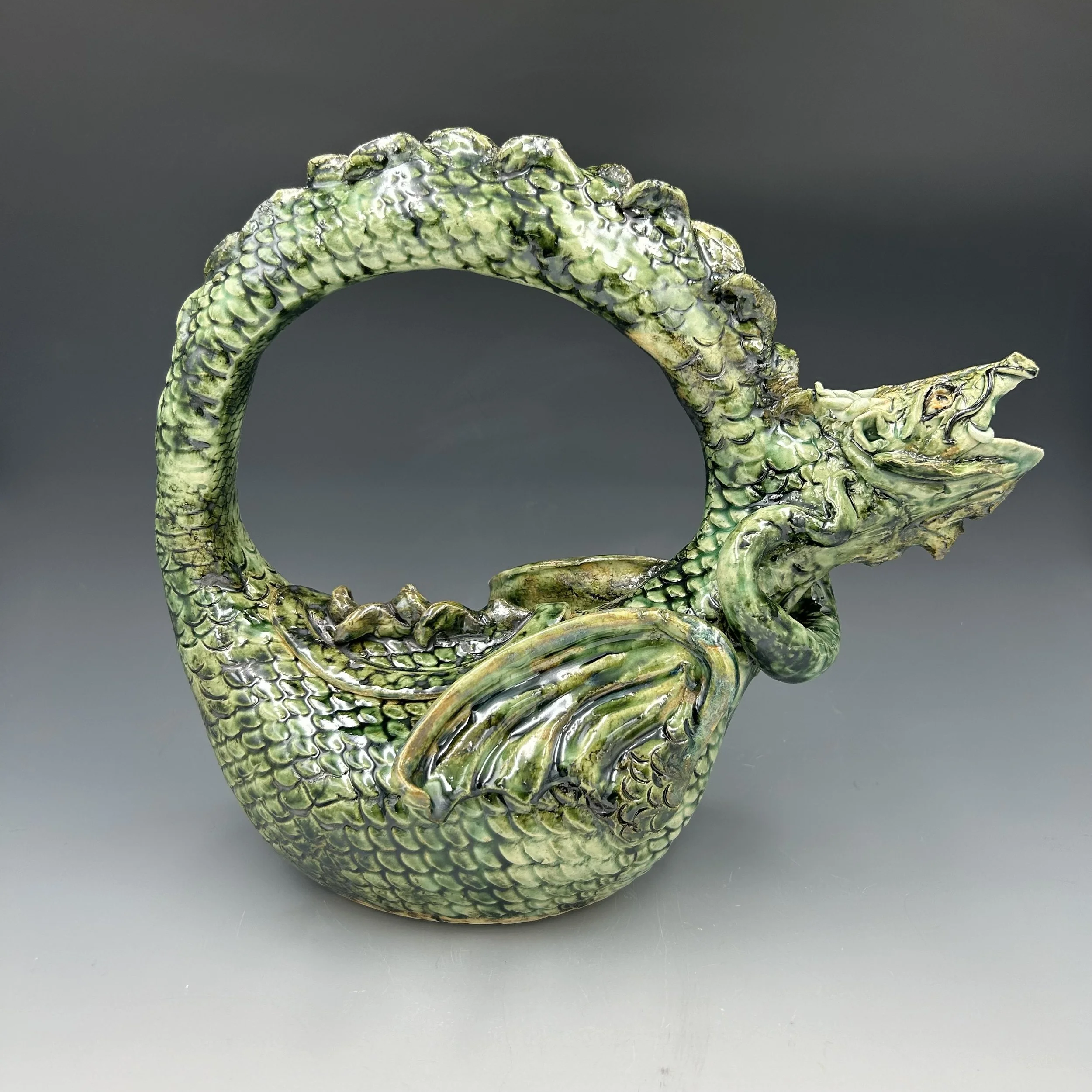A handmade ceramic teapot with a dragon shaped handle, and a body with scales and wings. The head and mouth form the spout.