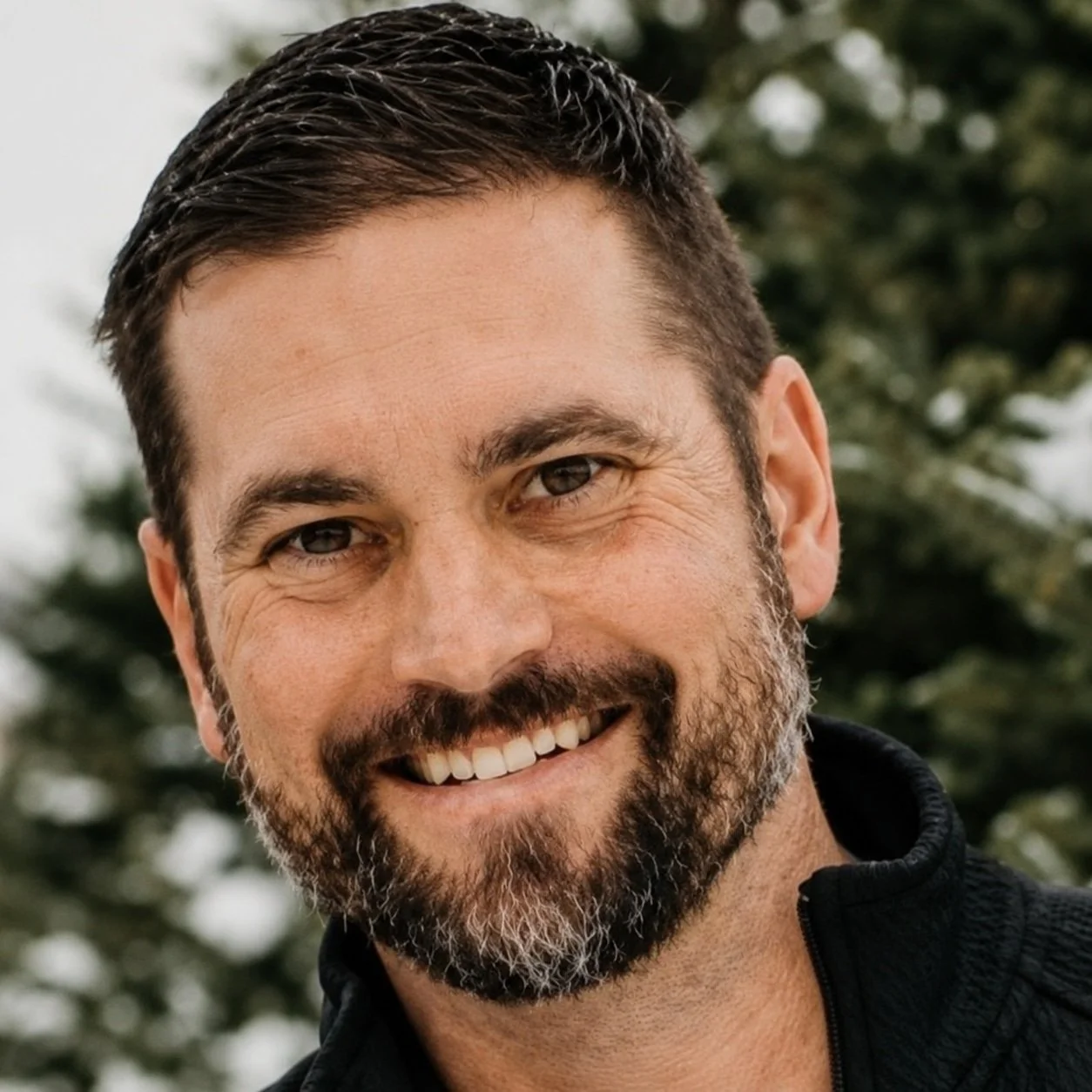 Close-up of a smiling man with dark hair and a beard, outdoors with blurred green trees in the background.