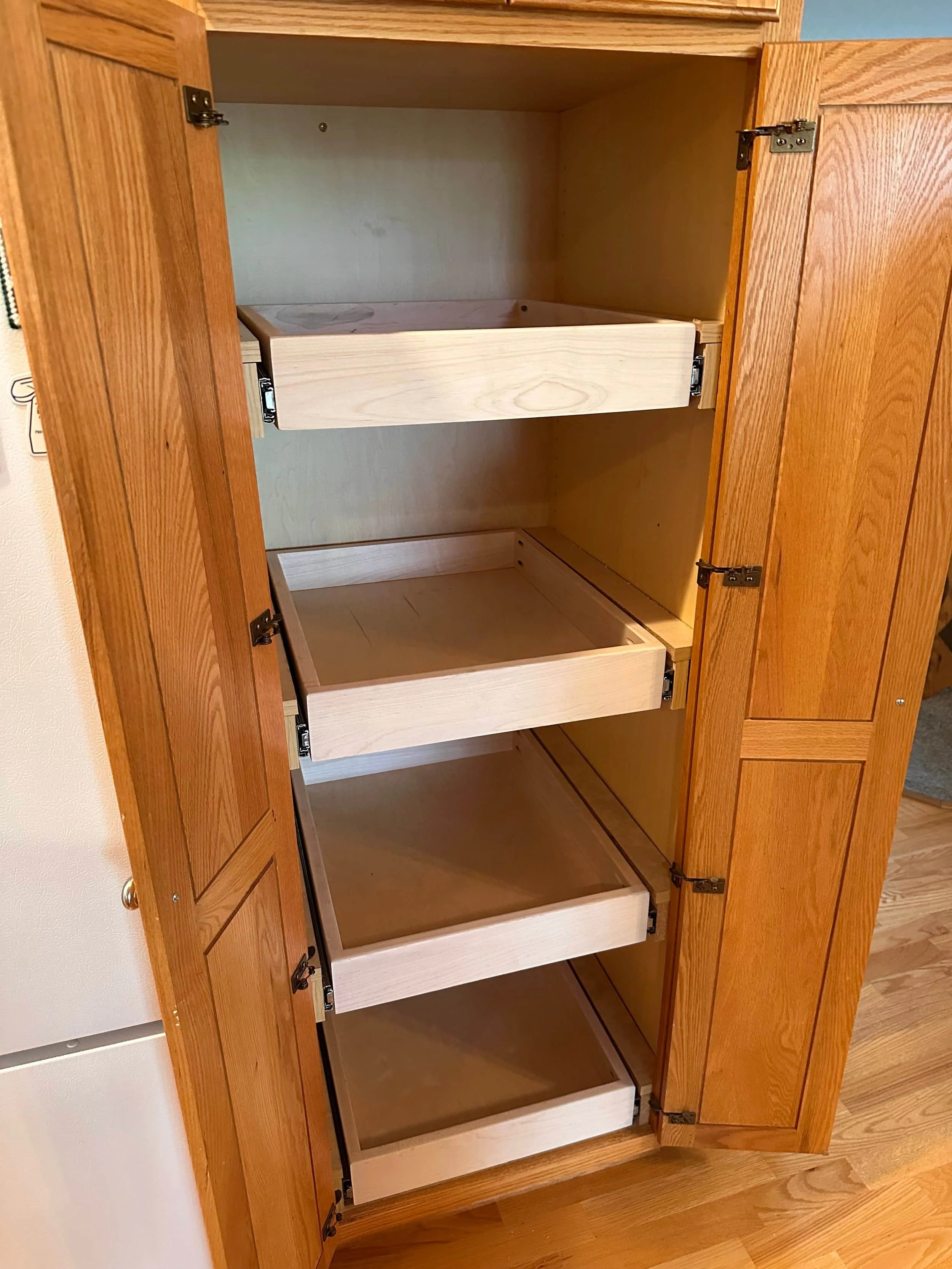 An open wooden cabinet with four empty drawers and two doors, showing hinges and handles, standing on a hardwood floor.