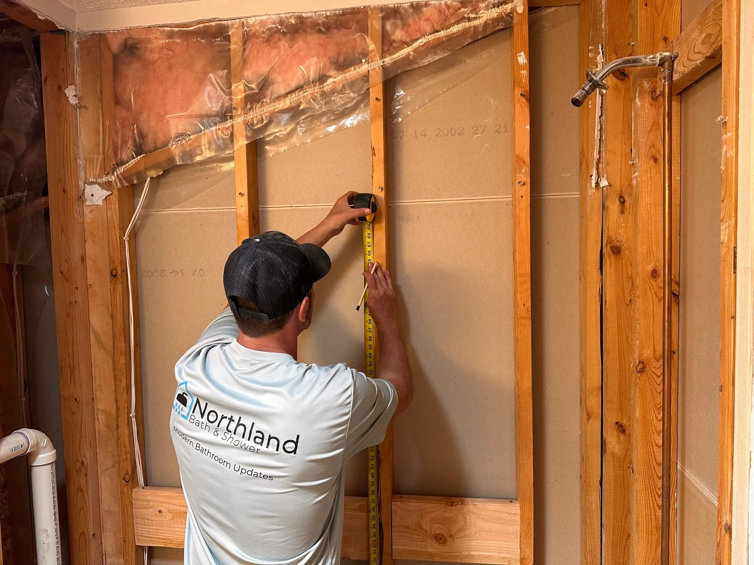 A man in a gray baseball cap and white shirt measuring the width of a wall with a tape measure. The wall has exposed wooden studs and some insulation and plastic sheeting reasons. The man is wearing a shirt with the logo and text of Northland Bath & Shower.