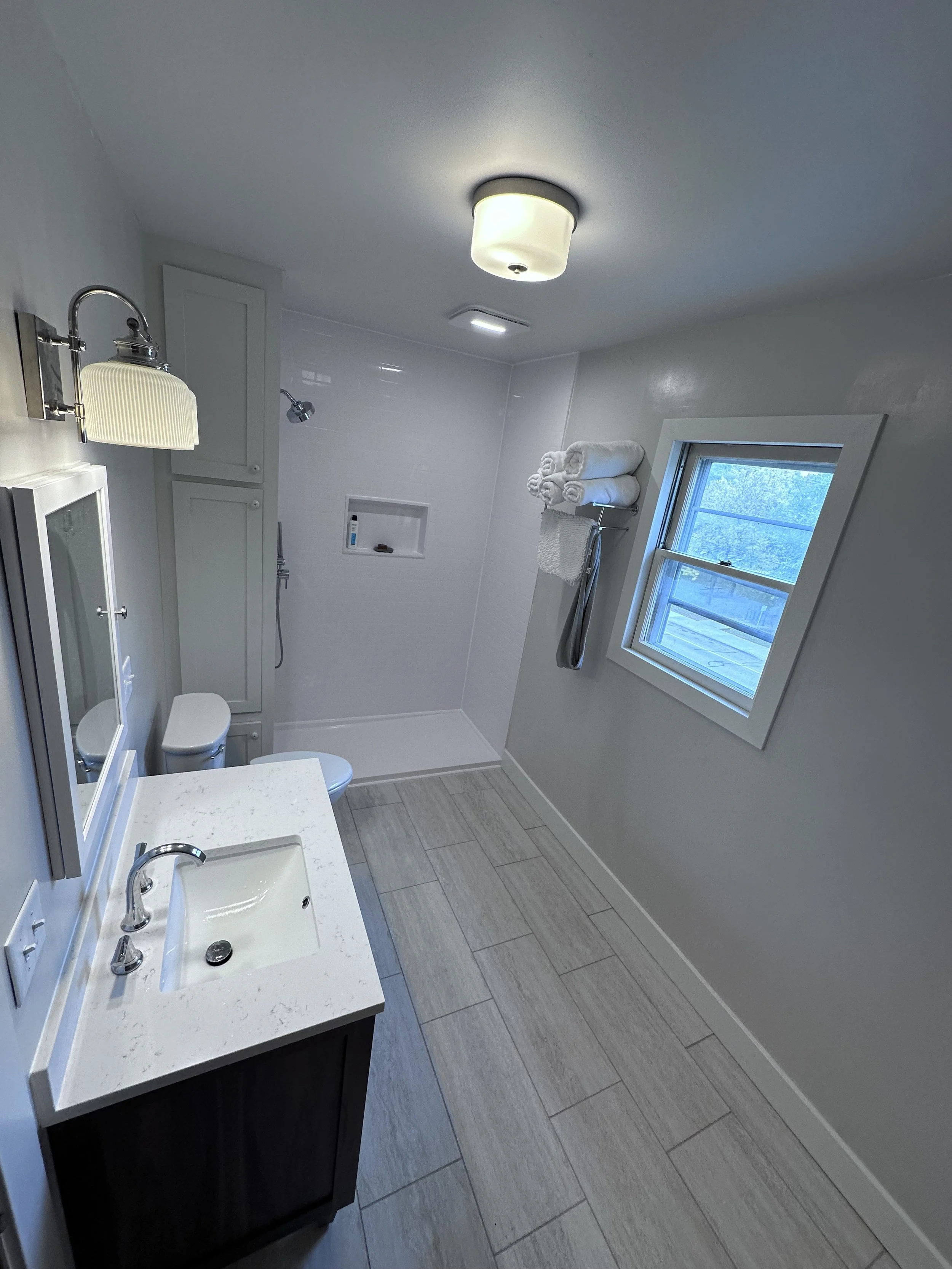A small, modern bathroom with a white sink vanity, a toilet, and a walk-in shower with a niche for toiletries. The walls are light-colored, and there is a window with white trim letting in natural light. Towels are stacked on a shelf above the shower