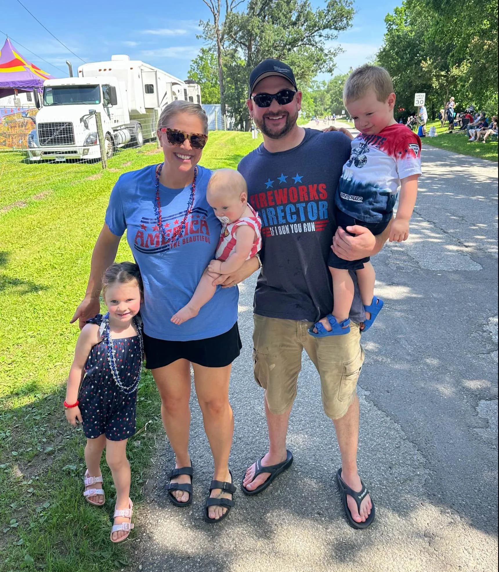 A family of five standing outdoors on a sunny day, smiling for the camera. The woman wears glasses and a blue shirt with patriotic text, holding a young girl in a navy dress with stars and stripes. The man wears sunglasses and a navy T-shirt with the text "Fireworks Director," holding a young boy in a red, white, and blue shirt. Older children are standing nearby, with the background showing a grassy area, trees, a parade, and people sitting on benches.