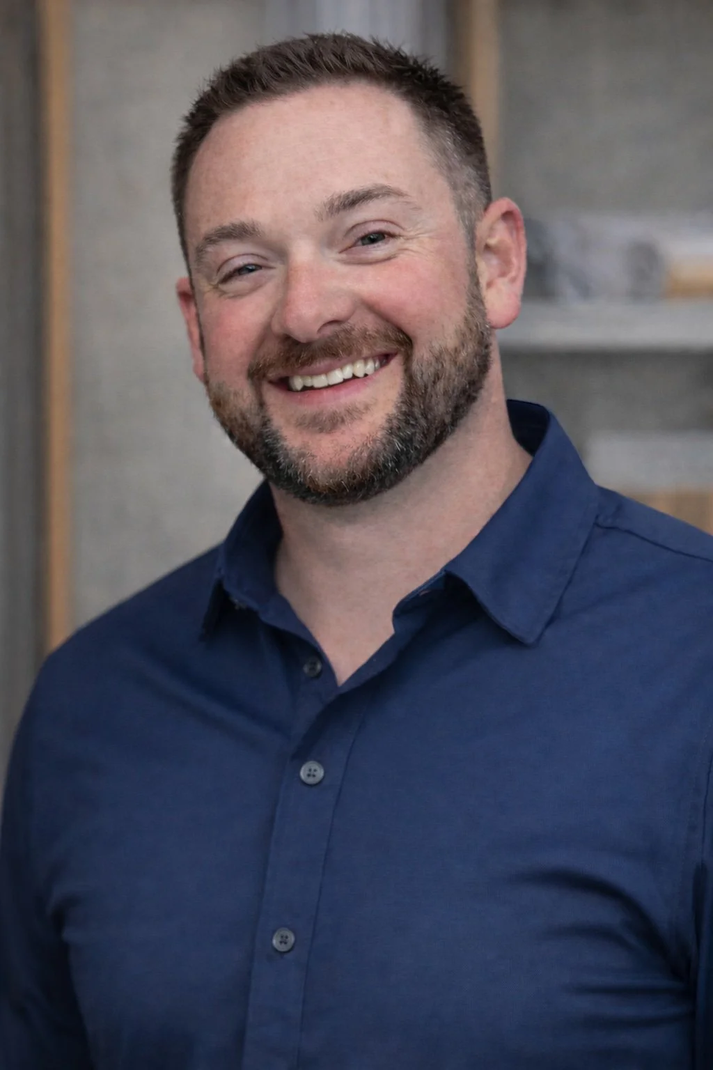 A smiling man with short brown hair and a beard, wearing a blue button-up shirt, standing indoors against a background with gray and wooden elements.