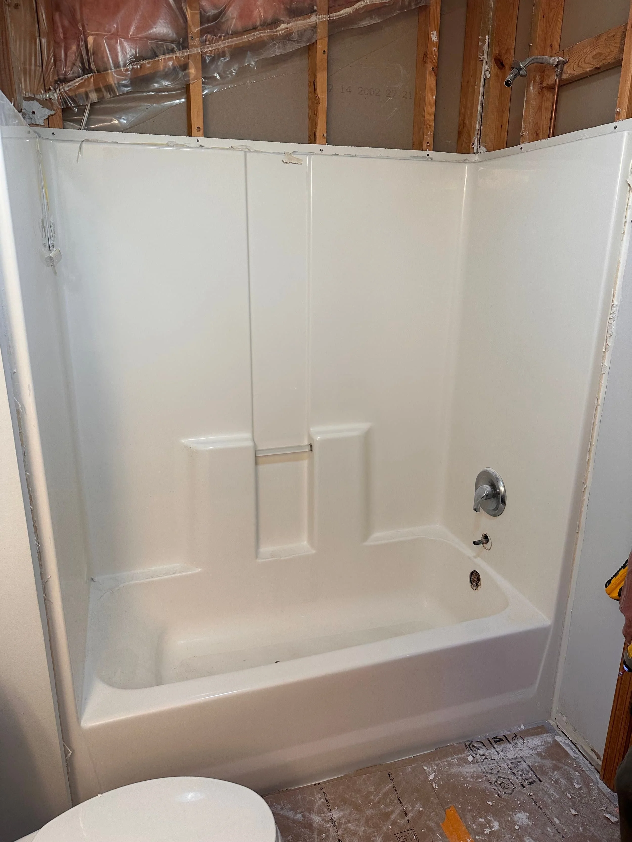 Bathroom shower area under renovation with a white acrylic shower stall, unfinished wall with exposed wooden studs, plumbing fixtures, and construction materials on the floor.