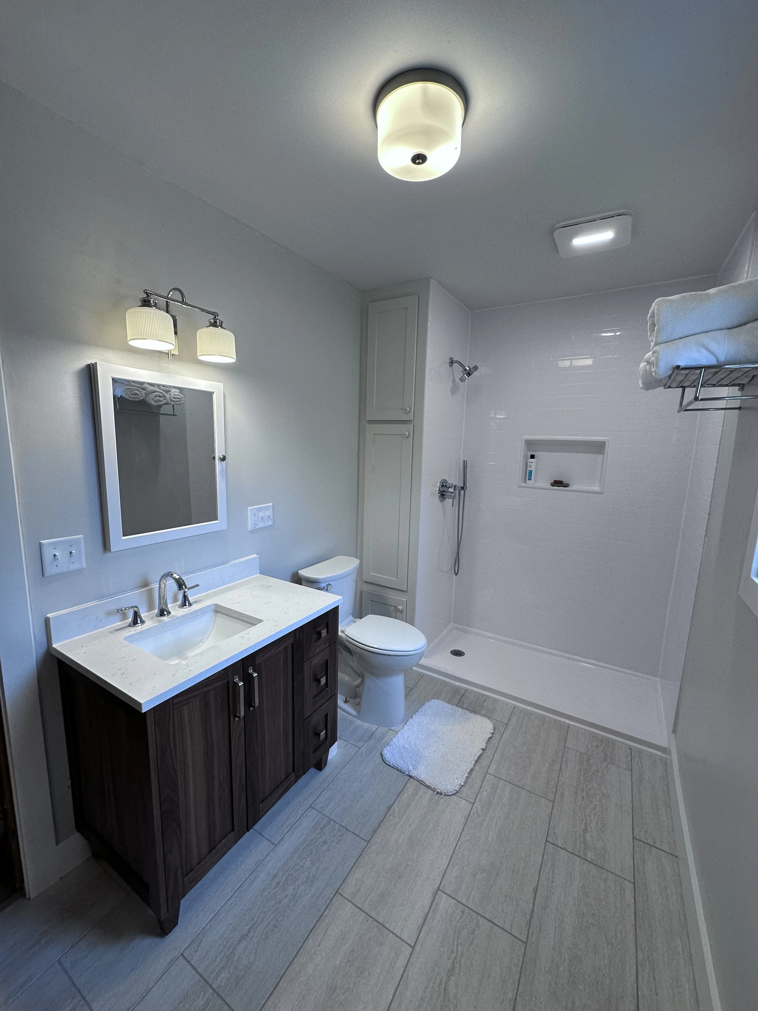 Bathroom with a dark wood vanity, white countertop, and mirror, white toilet, walk-in shower with glass door, towel rack with towels, ceiling light fixtures, and beige tiled floor.