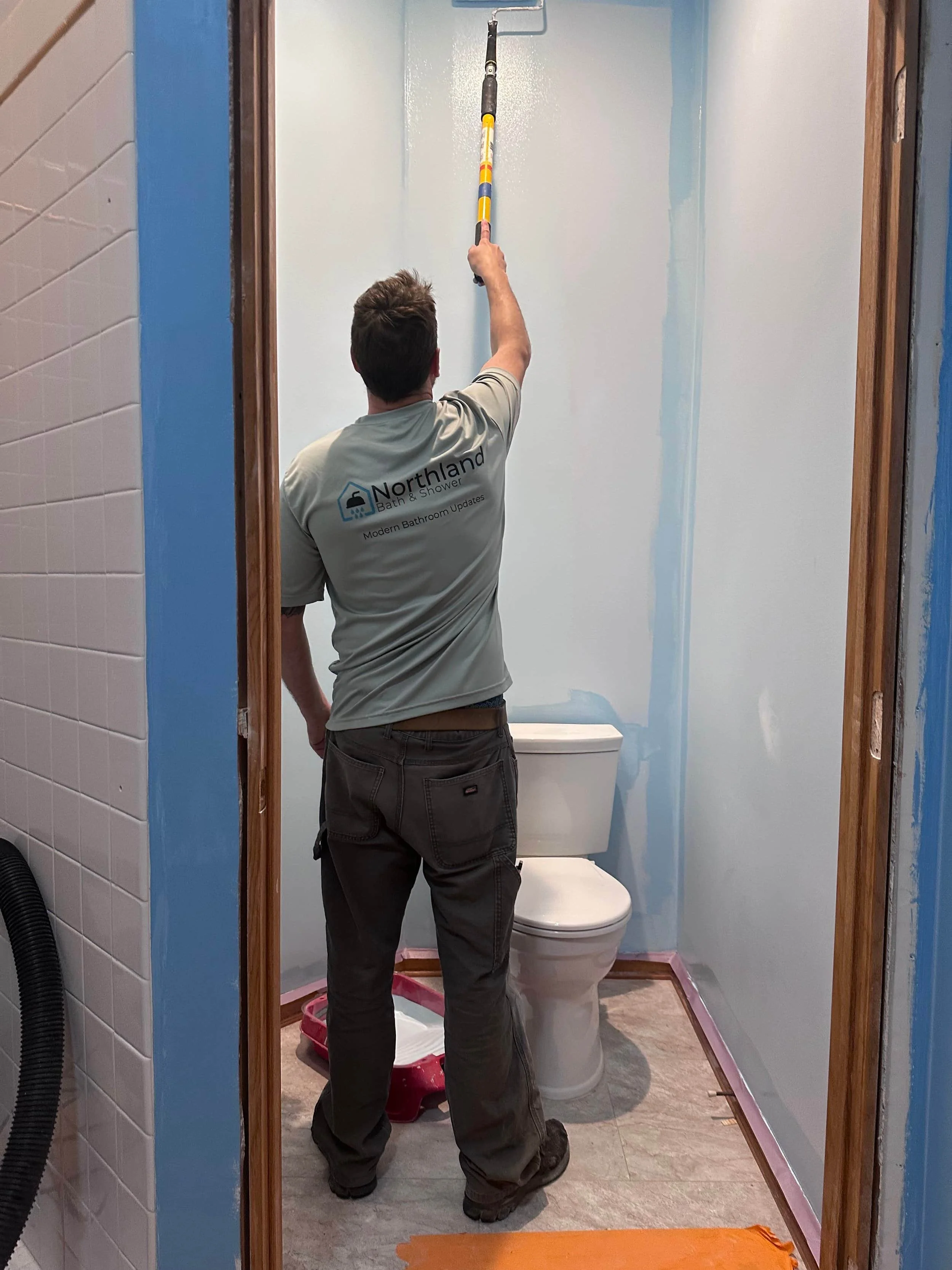 A man cleaning a bathroom wall with a long-handled roller brush. The bathroom has a toilet, and the walls are partially painted light blue. The man wears a gray shirt with the logo 'Northland Bath & Shower' on the back.