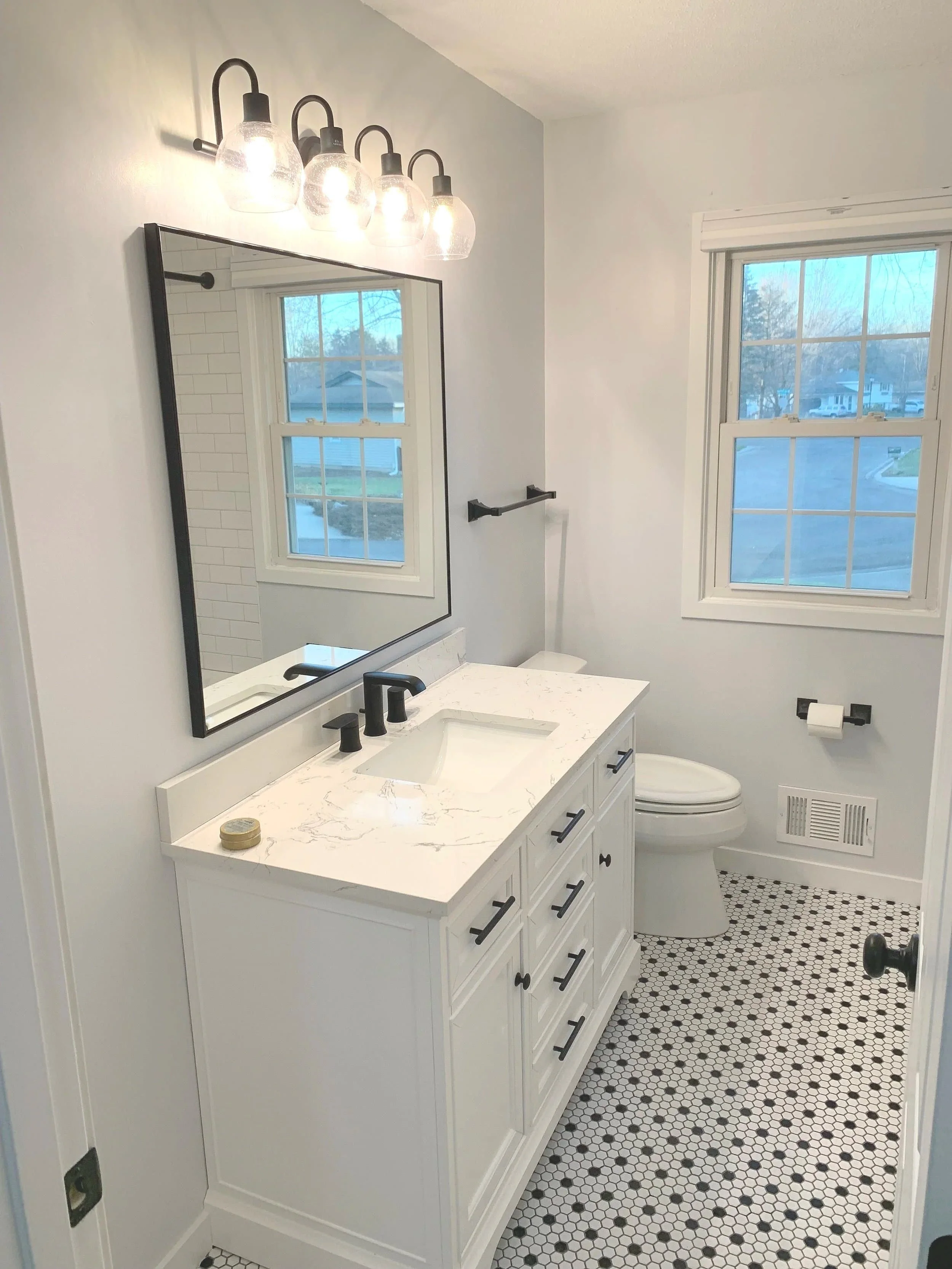 A clean bathroom featuring a white vanity with a marble countertop, black hardware, and a black faucet. Above the vanity is a large mirror and light fixture with four glass shades. There are two windows providing natural light, a toilet, a wall-mounted towel rack, a toilet paper holder, and a floor with black and white hexagonal tiles.