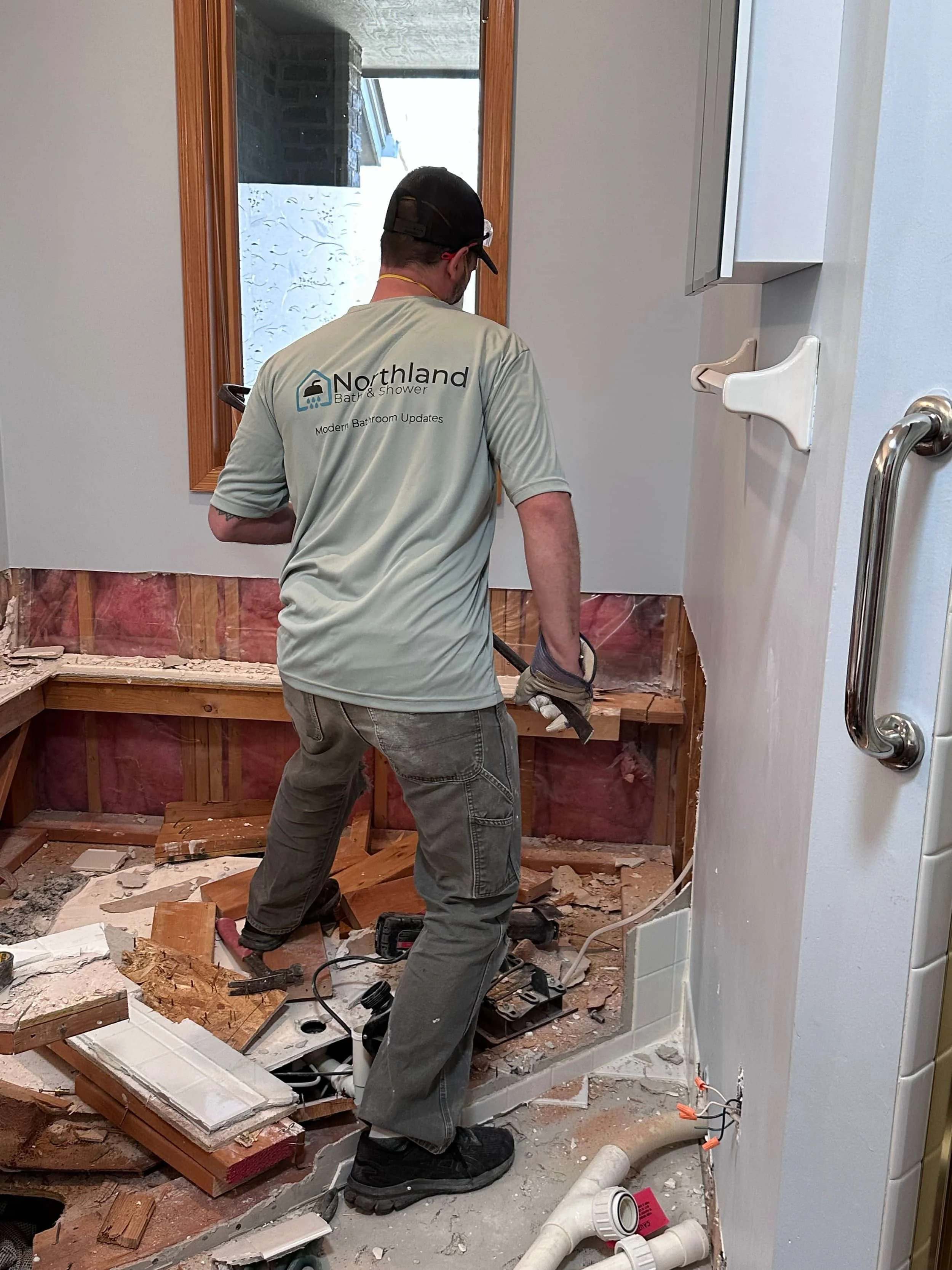 A worker in a beige shirt with 'Northland Bath & Shower' logo cleaning a bathroom under construction. The bathroom is partially demolished, with exposed wooden framing and debris on the floor. There is a window with a wooden frame and a white wall with a grab bar.