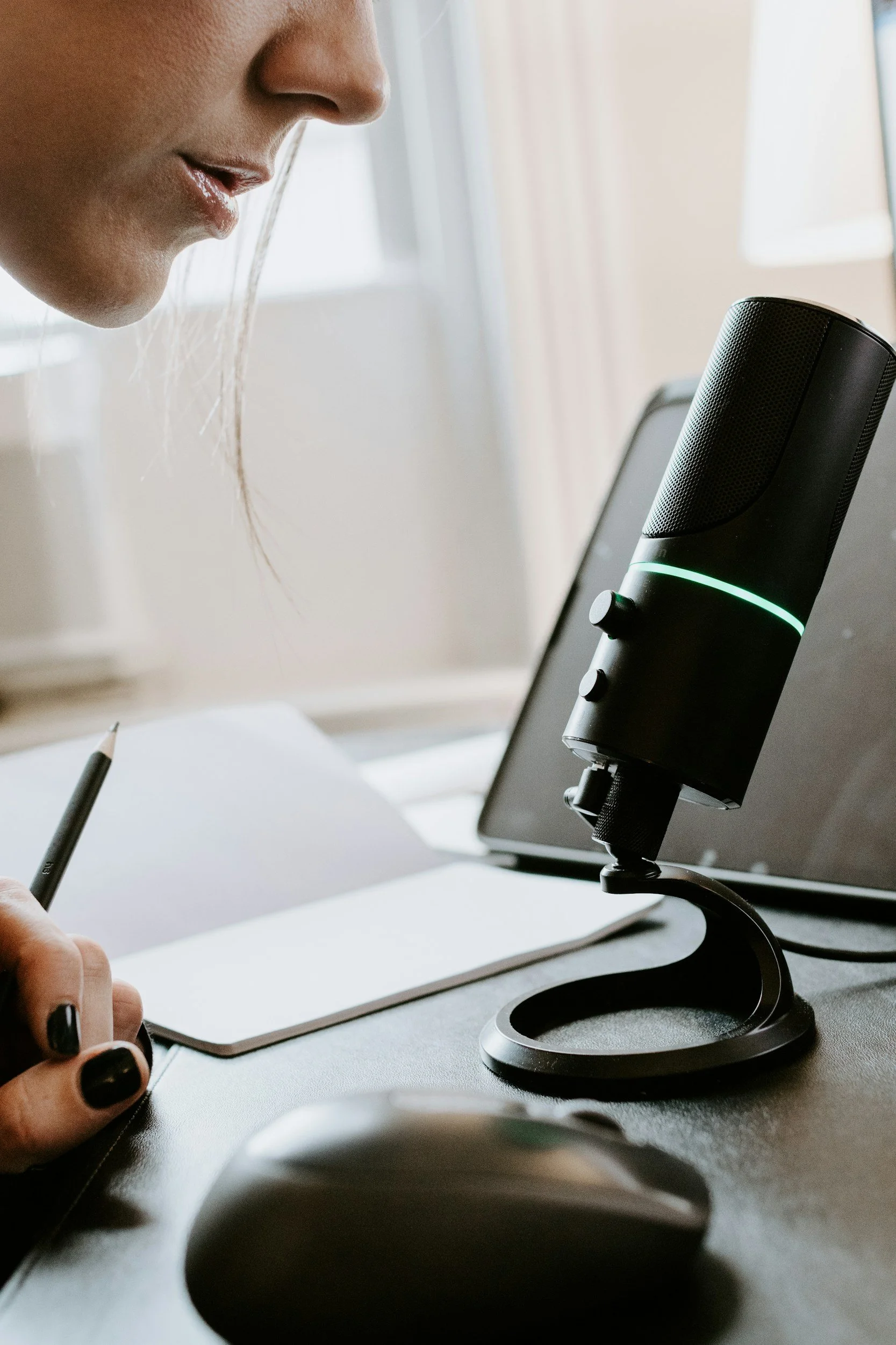 Close-up of a woman speaking into a black microphone with a green light, a laptop, notebook, pen, and mouse on a desk.